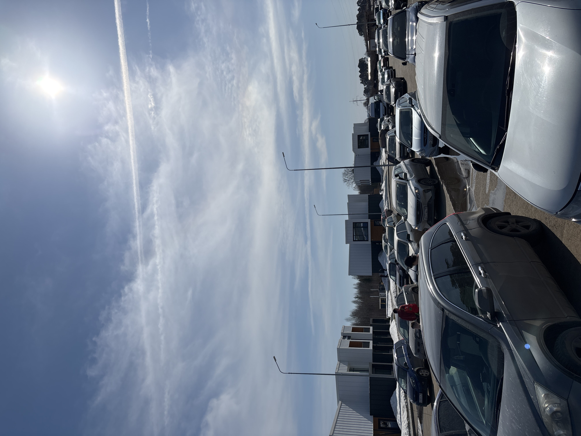 The image shows a parking lot filled with cars in Votkinsk, Russia. The sky is blue with scattered clouds and contrails. In the background are buildings and trees. A person wearing a red jacket is visible between the rows of cars. Streetlights are spaced throughout the parking lot. The image is taken during daylight hours under sunny conditions. There is some snow on the ground near the buildings.