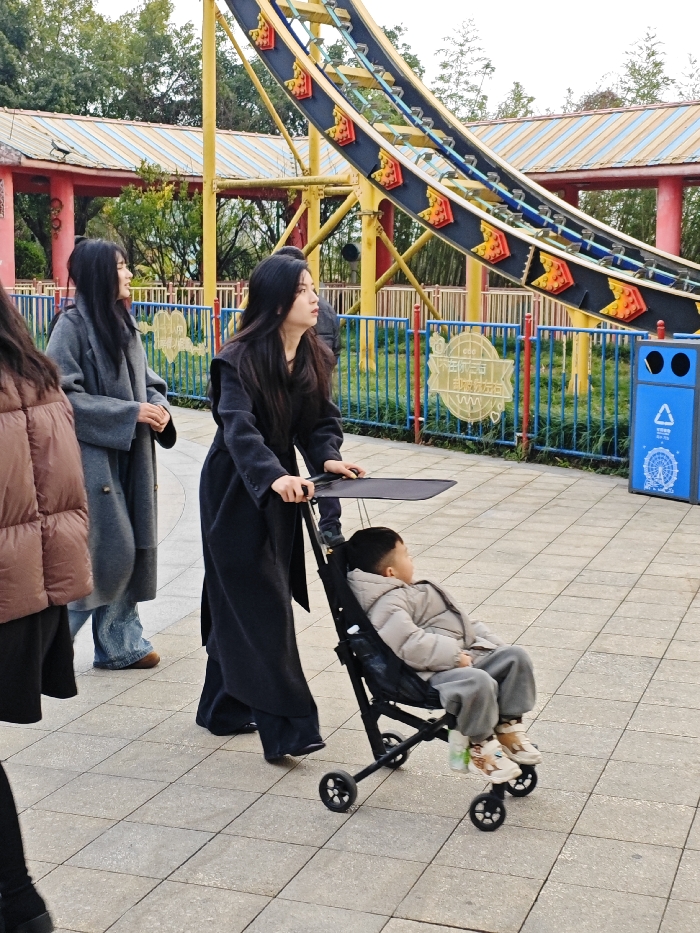 The image depicts an outdoor scene on a paved walkway within the Chongqing High-tech Zone, China. In the central foreground, a woman with long dark hair, wearing a dark long coat, pushes a black compact stroller. Seated in the stroller is a young child, wearing a light beige puffer jacket and grey pants, looking towards the right side of the frame. The woman pushing the stroller is looking upwards and to her right. To her left and slightly behind her stands another woman with long dark hair, dressed in a grey long coat and blue jeans, looking to her left. A person wearing a brown puffer jacket is partially visible on the far left.
In the mid-ground and background, a large yellow metal structure, consistent with an amusement park ride, extends upwards. It features a dark track segment embellished with red and orange arrow-shaped indicators. Behind this structure, green trees and red-colored buildings with striped roofs (yellow and blue) are visible. A blue metal fence with vertical bars and a circular yellow sign featuring Chinese characters runs along the perimeter of a grassy area to the right. A blue recycling bin with multiple compartments and a graphic of a Ferris wheel is positioned on the far right. The ground is paved with light-colored square tiles.