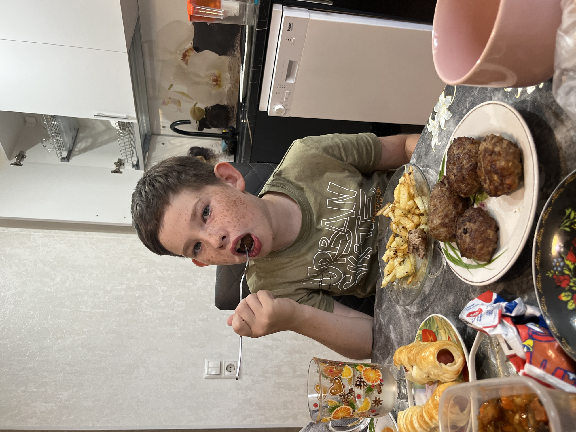 A young boy with freckles is eating a meal at a table. He is wearing a green t-shirt with the words "URBAN SKATE" on it. He is using a fork to eat what appears to be a meatball. On the table, there is a glass mug with a festive design, a plate of french fries and meatballs, a plate with two sausage rolls, a container of food, and a pink mug. In the background, there is a kitchen counter with cabinets above it. A dishwasher is visible on the right side of the image. The location is the city of Chaikovsky, Russia.