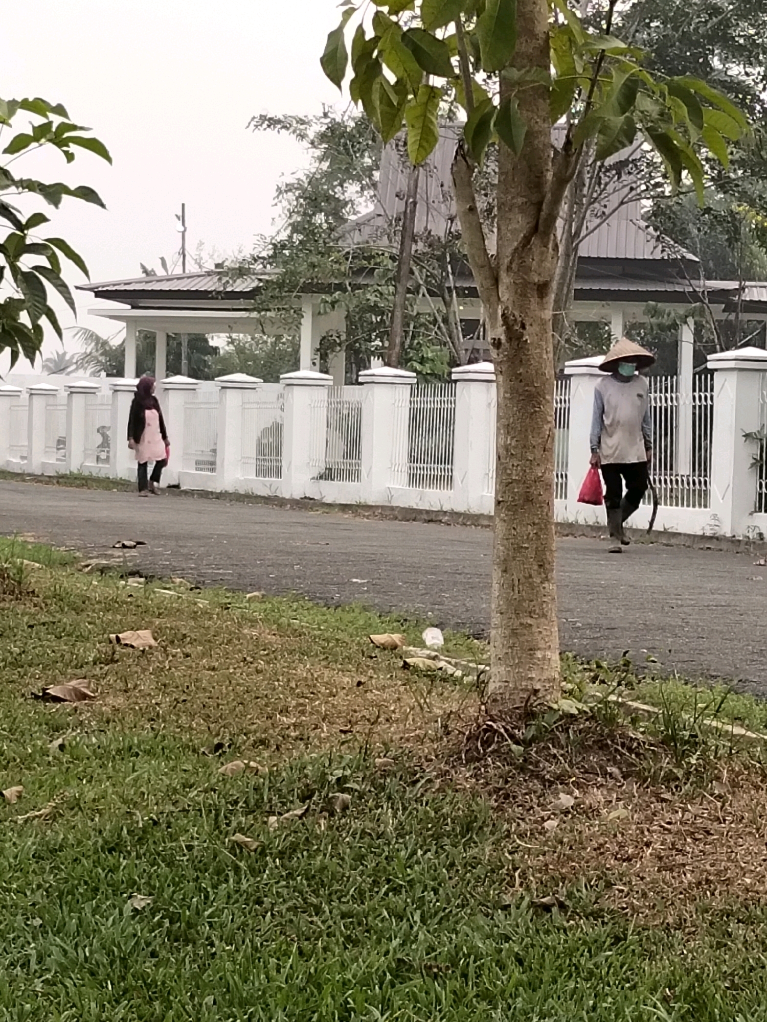 The image depicts a street scene in Sungai Raya, Indonesia, with two individuals walking along a paved road. A white decorative fence with vertical bars and a capstone runs along the right side of the road, bordering a grassy area and a building with a silver roof. In the foreground, a tree trunk is prominent, with green foliage visible at the top.
On the right side of the road, a person wearing a conical hat and a blue mask walks away from the camera, carrying a red plastic bag in their left hand and holding a dark object, possibly a tool, in their right hand. They are dressed in a light-colored long-sleeved shirt and dark trousers, with dark boots.
On the left side of the road, further in the distance, another person walks towards the camera, also moving away from the building. This individual wears a dark head covering, a light pink apron over a dark long-sleeved garment, and dark trousers.
The environment appears to be outdoors, with a slightly overcast sky and some visible greenery, including grass and trees. The overall impression is of a rural or semi-urban setting in Indonesia.
