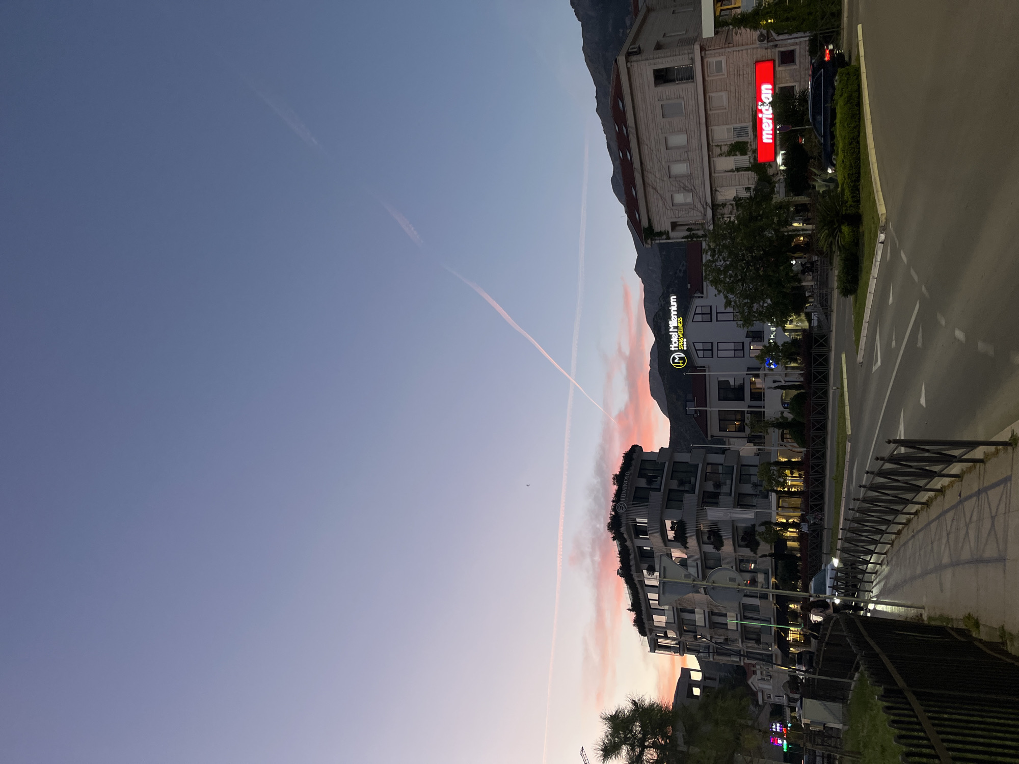 The image captures an urban street scene at dusk in Budva, Montenegro, with buildings and mountains set against a colorful sky. The sky, occupying much of the left side, transitions from deep blue at the top to lighter blues, purples, and pinks closer to the horizon, indicative of sunset. Two contrails, one long and faint, the other shorter and pink-tinted, streak across the sky. A small, dark shape, possibly an airplane, is faintly visible in the upper sky.
On the right, a series of multi-story buildings line a street. Artificial lights are visible within windows and on building facades, reinforcing the evening setting. Notable signs include a prominent red sign with white text that reads "meridian" on a light-colored building, and another sign for "Hotel Millenium Spa & Wellness" with an 'LM' logo on a white building. Further along, a modern building features the text "LUMINOSO".
A paved road runs along the far right, with a sidewalk separated by a dark, decorative metal fence in the foreground. Shadows from the fence are cast on the sidewalk. Greenery, including trees and bushes, is interspersed along the street. Mountains rise in the background, silhouetted against the fading light of the sky. Several parked or moving vehicles are scattered along the street. The overall atmosphere is calm and serene, capturing the transition from day to night in an urban setting. No explicit human activity is visible, but the presence of urban infrastructure implies city life. The weather appears clear with minimal cloud cover.