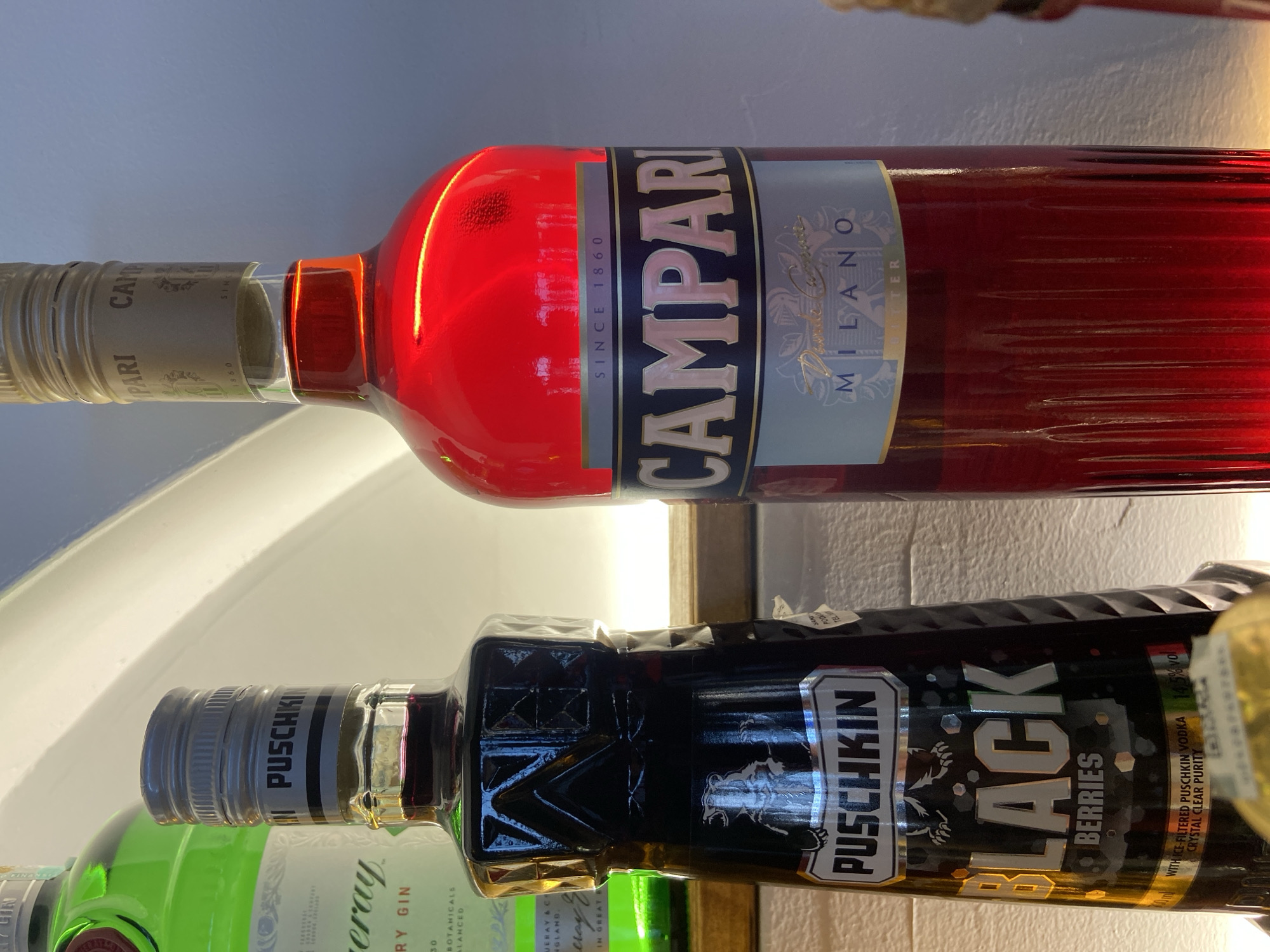 The image shows three alcohol bottles on a shelf. The top bottle is Campari, a red-colored aperitif with a label that reads "CAMPARI MILANO SINCE 1860." Below the Campari bottle is a bottle of Puschkin Black Berries vodka with a black label featuring the Puschkin logo and the words "BLACK BERRIES." Part of another green liquor bottle is visible at the bottom left, with "Finest Dry Gin" written on it. The shelf is white, and the lighting is bright. There is no information about the location in Petrovac centar, Montenegro or other elements.