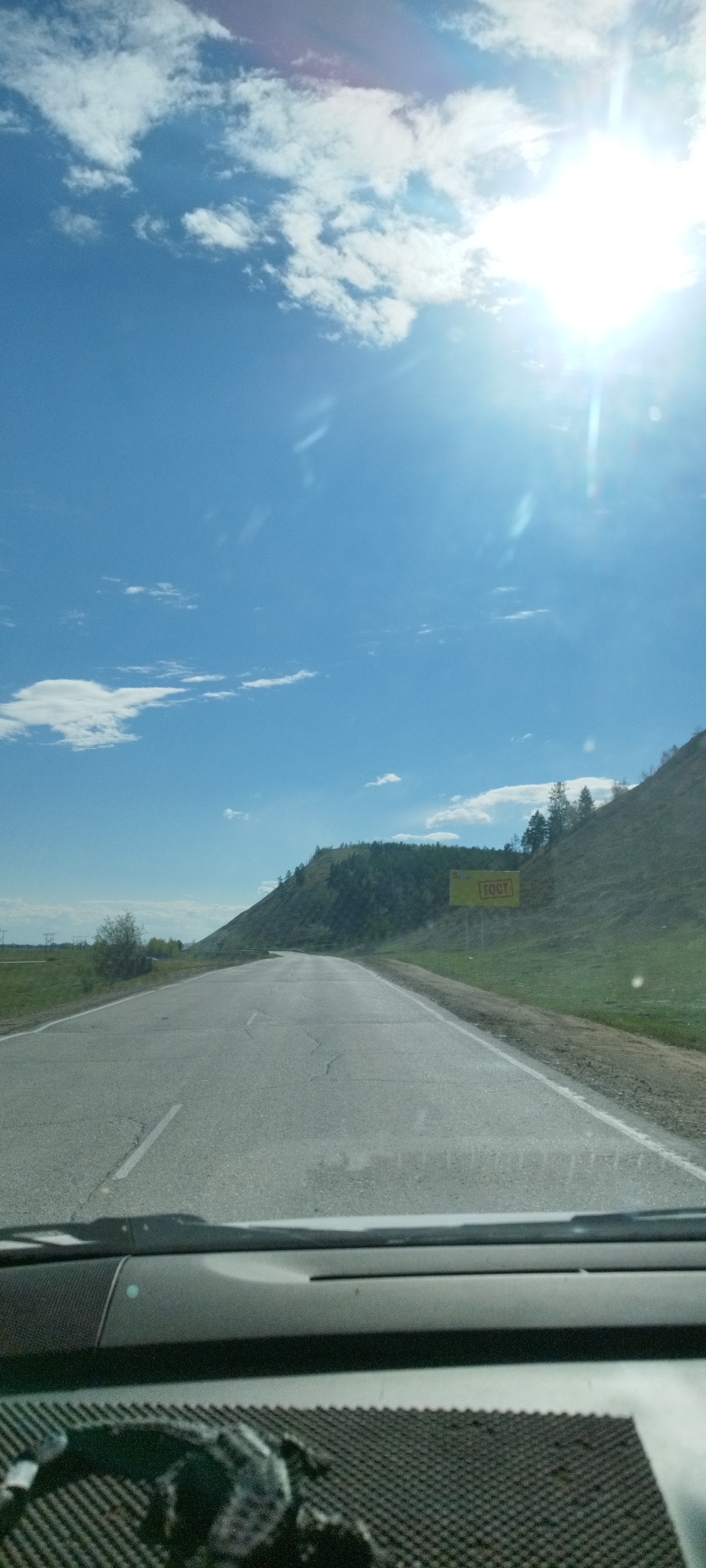 The image depicts a view from inside a vehicle, looking out onto a paved road. The road leads towards a small hill covered in vegetation. A large yellow sign with the word "ГОСТ" in Russian is visible on the right side of the road, near the base of the hill. The sky is blue with scattered white clouds and bright sunlight. The dashboard of the vehicle is visible at the bottom of the frame. The location is identified as Tekhtyur, Russia.
