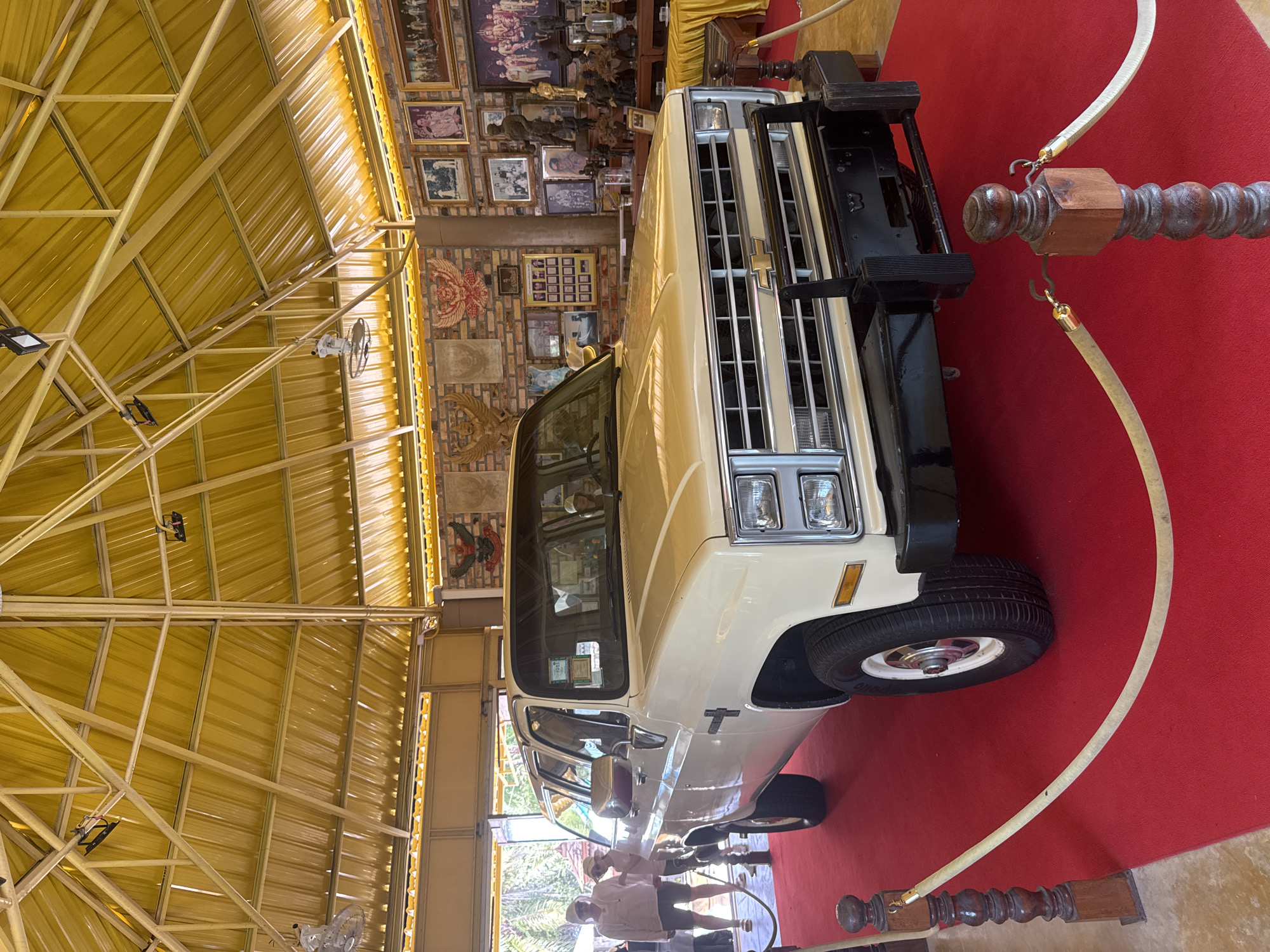 Captured indoors in Ban Khlong Khian บ้านคลองเคียน, Thailand, the image features a cream-colored Chevrolet pickup truck prominently displayed on a red carpet. The front of the truck, with its black bumper, chrome grille, and headlights, dominates the foreground. The truck is positioned close to a roped-off barrier. The environment consists of a building with a yellow corrugated metal roof structure supported by metal beams. The back wall is adorned with multiple framed pictures and decorative items. People are visible in the background near a doorway.