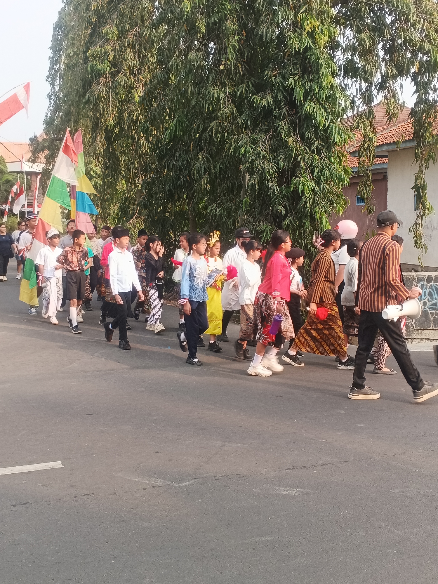 A group of children and young people are participating in a parade on a paved road in Sokanegara, Indonesia. They are walking from left to right in the frame, some holding flags. Several individuals are wearing traditional Indonesian clothing, including batik skirts and shirts. One person in the foreground on the right is holding a megaphone. The background features lush green trees and terracotta-tiled rooftops of houses, suggesting a residential area. The sky is visible at the top of the image and appears to be a bright, clear day. The road has some white markings. The overall atmosphere appears festive and celebratory.