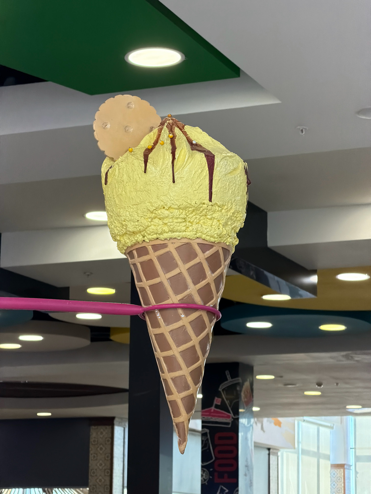 The image shows a large, artificial ice cream cone sculpture hanging from the ceiling. The ice cream is yellow with chocolate drizzle and what appear to be sprinkles. A round, flat cookie is perched on top of the ice cream. The cone is brown and textured like a waffle cone. A bright pink support rod is wrapped around the cone. The background consists of a ceiling with a green panel and circular lights, as well as other decorative ceiling elements in yellow and blue. The overall setting appears to be an indoor public space, possibly a shopping mall or restaurant, given the lighting and architectural features. No people are visible in the image. The location is specified as Pavlodar, Kazakhstan, though the image itself does not contain any visual cues to confirm this specific location beyond what is provided in the prompt.