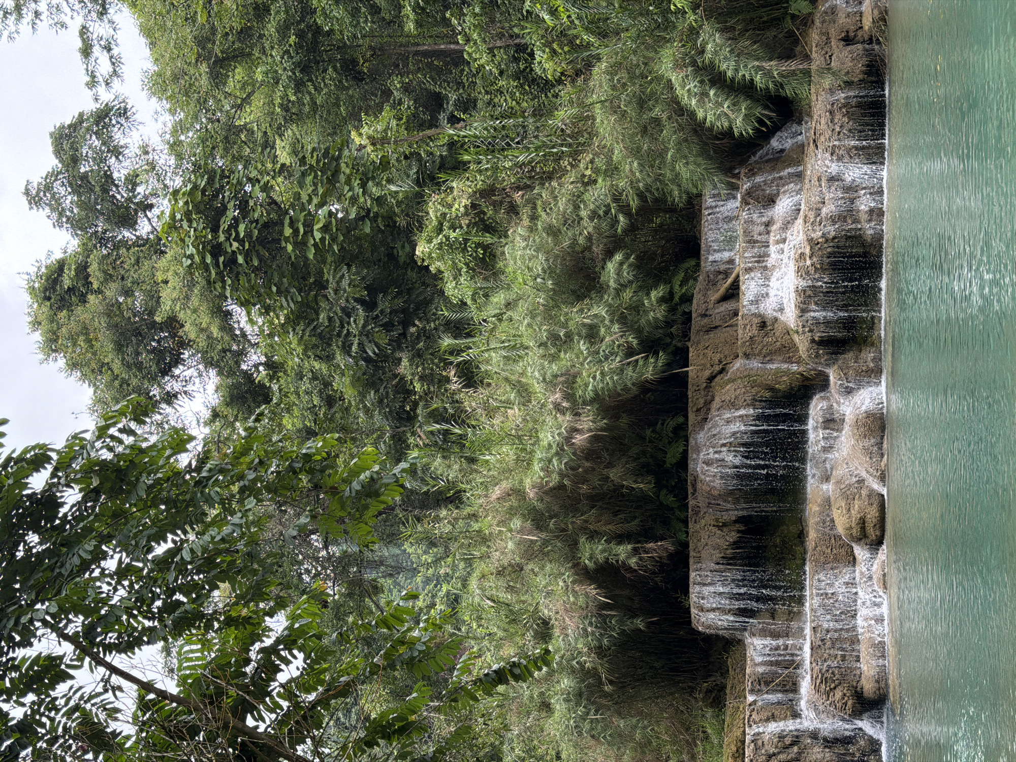 A cascading waterfall flows over multi-tiered, eroded rock formations into a pool of turquoise water. Lush green vegetation, including dense bamboo and tall trees, surrounds the waterfall and forms a thick jungle backdrop under a cloudy sky. The water appears clear and inviting. The scene is a natural landscape within the city of Thapene, Laos, highlighting the beauty of its natural attractions.