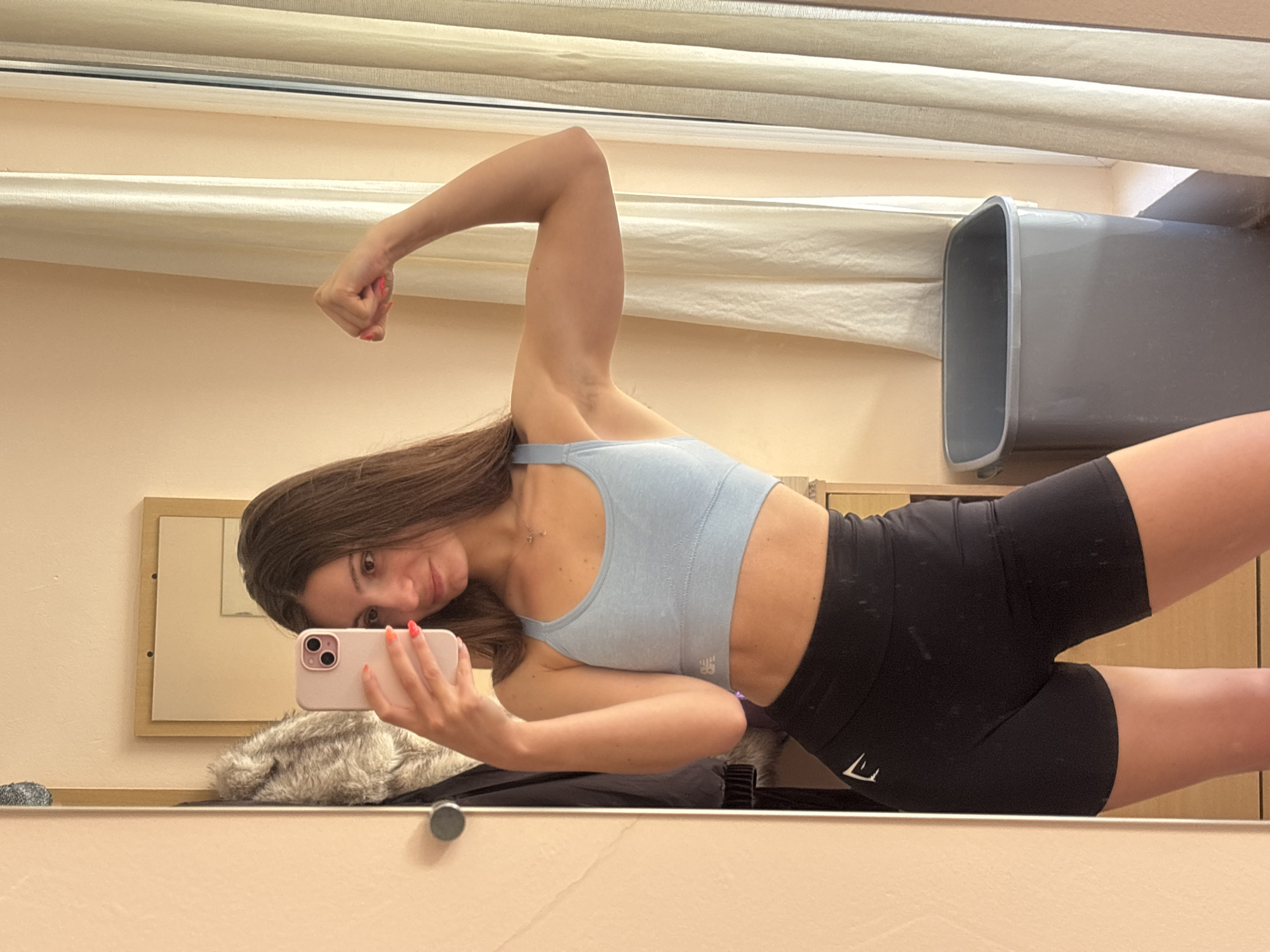 A woman is posing in front of a mirror, flexing her right bicep and taking a selfie with a pink smartphone. She is wearing a light blue sports bra and black shorts. The background shows a beige wall, a window with drawn curtains, a gray trash can, and a framed mirror. The location appears to be indoors, potentially a bathroom or dressing room. The city of Varna, Bulgaria is not directly discernible from the image.