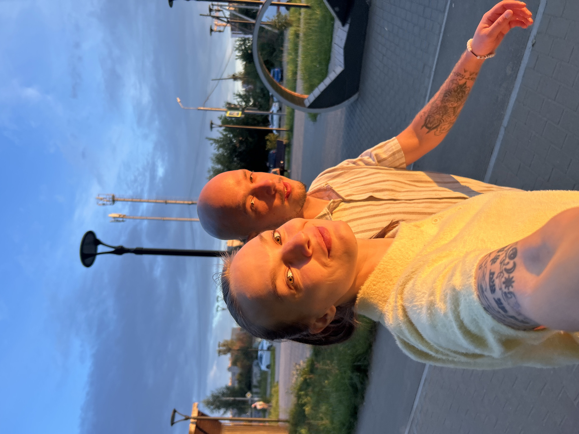 This is a selfie taken outdoors in the city of Chaikovsky, Russia, during sunset. The warm, golden light of the setting sun illuminates the faces of a man and a woman. The woman is in the foreground, smiling at the camera, with her arm extended as if holding the phone. She has a ponytail and is wearing a yellow, fuzzy sweater. Her left forearm is visible, showing a tattoo.
The man is standing next to her, also looking towards the camera, with a slight smile. He is bald and wearing a striped shirt. His right arm is visible, and it also has tattoos.
The background shows a street scene with cars, trees, and streetlights. There are also tall cell towers in the distance. The sky is a mix of blue and gray clouds. The ground is paved with bricks. There is a modern-looking bench or seating area in the mid-ground. The overall impression is that of a couple enjoying an evening walk as the day ends.
