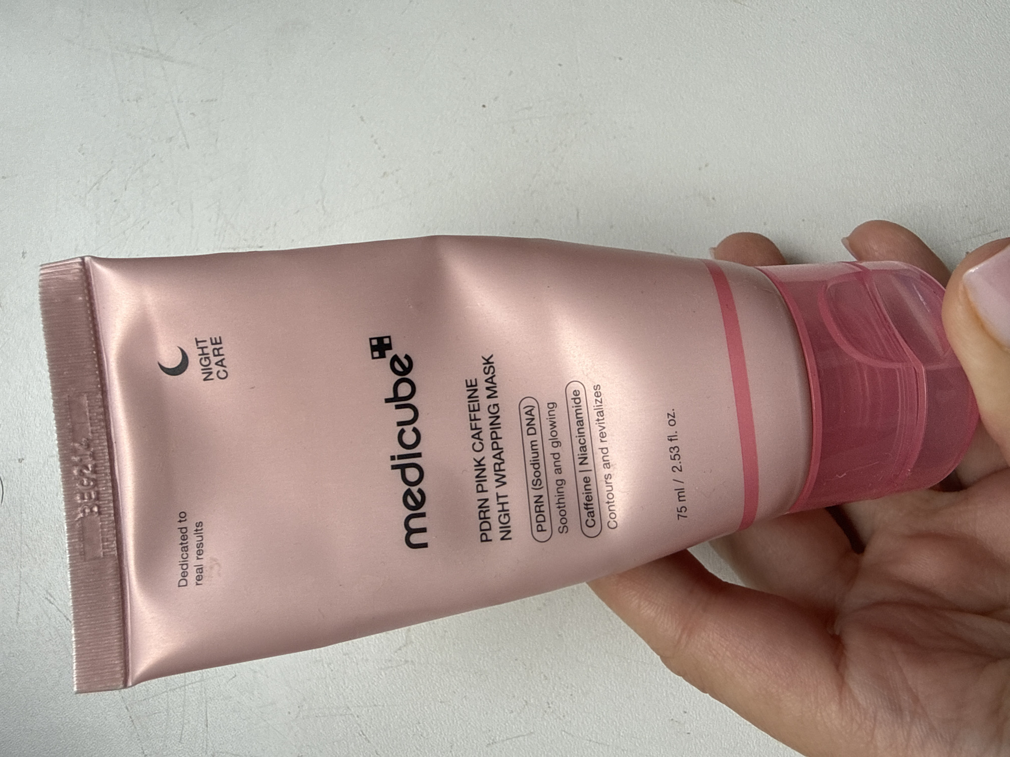 A person's hand, with light-colored skin and light pink nail polish, holds a pink tube of Medicube PDRN Pink Caffeine Night Wrapping Mask. The tube is made of a light pink, somewhat shiny material. Text on the tube includes "Dedicated to real results," "medicube," "PDRN PINK CAFFEINE NIGHT WRAPPING MASK," and "75 ml / 2.53 fl. oz.". A small crescent moon symbol and the words "NIGHT CARE" are also visible. The background appears to be a light-colored, textured surface, possibly a desk or countertop. There is no indication of the city of Okhtyrka, Ukraine, in the image or its metadata.