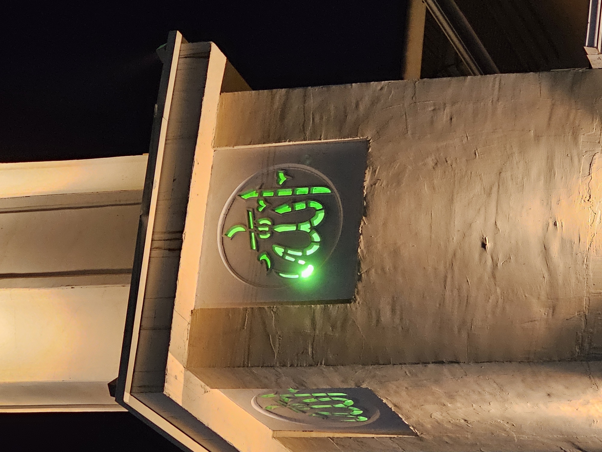 The image captures a close-up of a section of a minaret at night, illuminated by warm-toned artificial lights and featuring a prominent green neon Arabic calligraphy design. The minaret appears to be constructed from textured concrete or stucco, with a lighter colored, possibly stone or painted, decorative element near its top. The calligraphy is circular and appears to be the word "Allah" in Arabic script, rendered in a stylized, glowing green neon outline. There are two such circular designs visible, one more centrally placed and brightly lit, and another partially visible to the left, also in green neon. The background is dark, indicating nighttime, with a black sky and the suggestion of further structures or the continuation of the building beyond the frame. The lighting creates strong shadows and highlights on the textured surface of the minaret, emphasizing its architectural details. The scene is devoid of people and focuses solely on the architectural elements and the decorative calligraphy. The location is specified as Özgön, Kyrgyzstan.