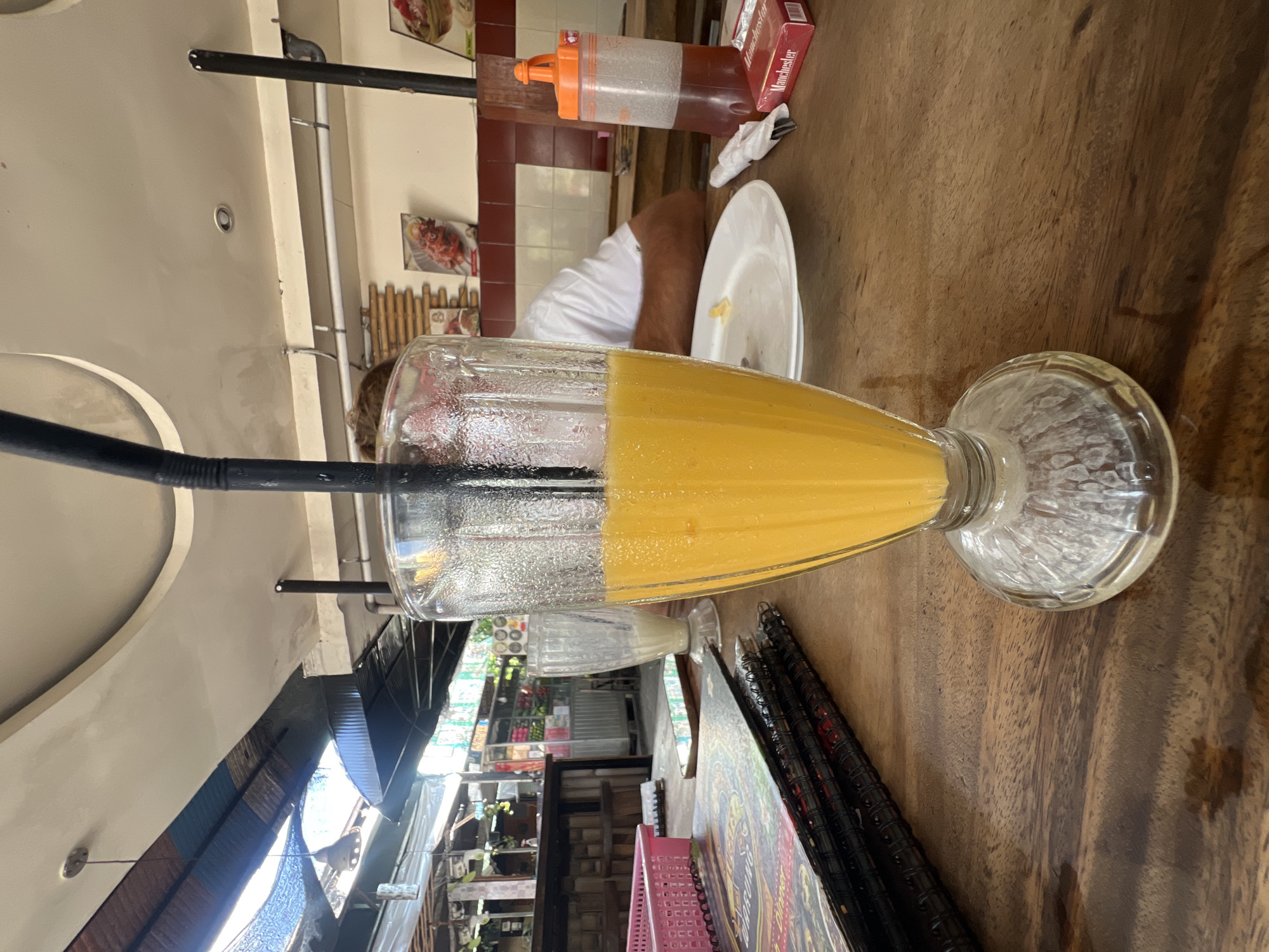 The image shows a table at an outdoor restaurant in Kuta, Indonesia. A tall glass filled with orange juice is in the foreground, with a black straw inserted. To the left of the juice glass are several menus stacked on the table. In the background, a person wearing a white shirt is seated, and there are various decorations and restaurant elements, including a condiment bottle, a plate with food remnants, and cigarette packs. The environment appears to be casual and relaxed, typical of a beachside eatery in Kuta.