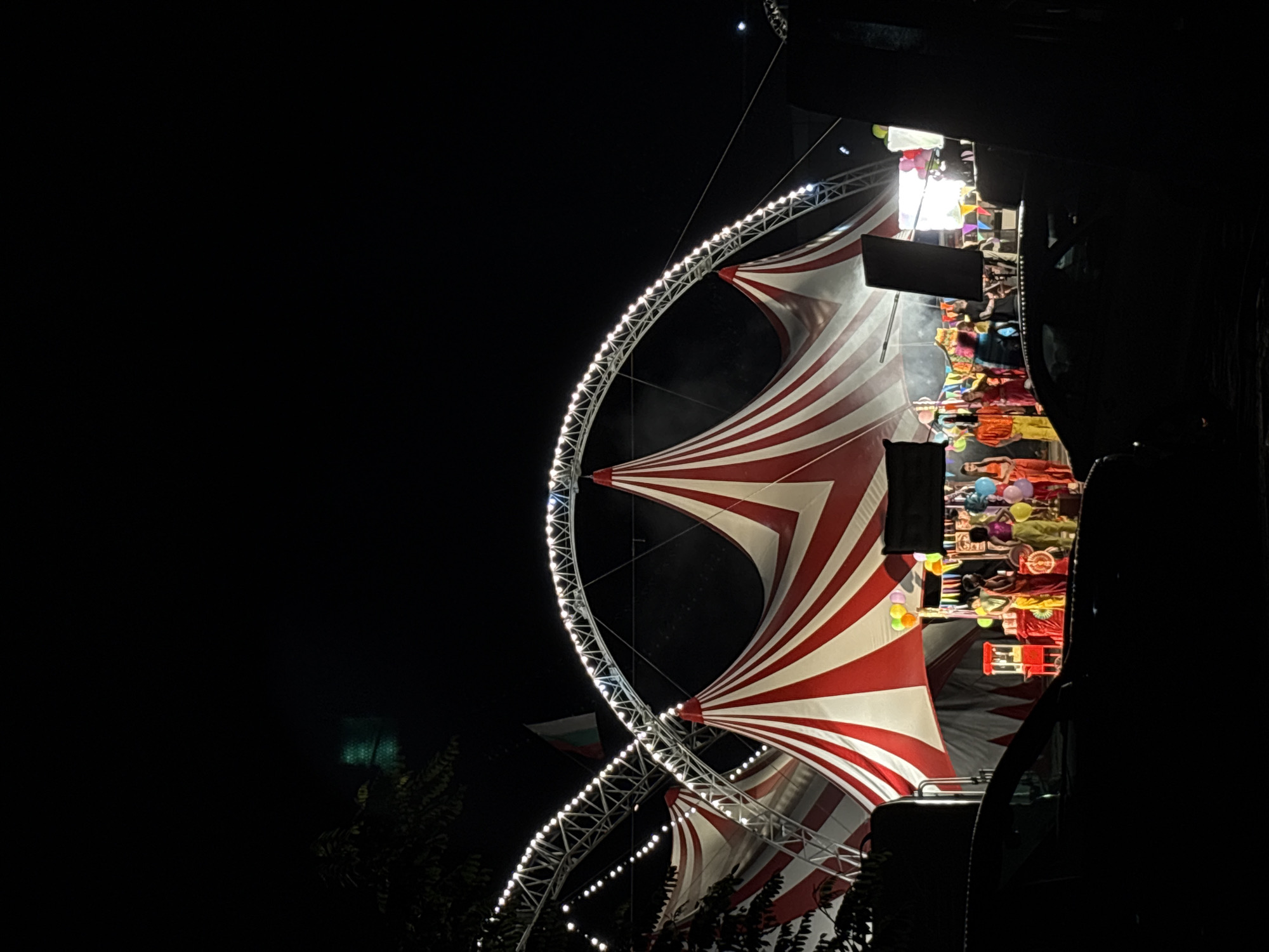 The image depicts a vibrant night scene at an outdoor event, characterized by festive lighting and colorful structures. The central focus is a large, elaborate tent or canopy featuring prominent red and white stripes that create a dynamic, undulating pattern.
To the left, a large curved metal truss, illuminated by numerous small, bright lights, arches upwards into the dark night sky. This structure appears to be an integral part of the event's setup, possibly supporting the tent or serving as a decorative entrance. Below this, some dark green foliage suggests the presence of trees. A Bulgarian flag, identified by its white, green, and red horizontal stripes, is partially visible in the upper left section, confirming the event's location in Bulgaria, specifically Plovdiv.
The right side of the image is brightly illuminated, revealing a bustling area that appears to be a stage, performance space, or a series of market stalls. Several people are visible here, adorned in colorful clothing, though their individual features are indistinct due to the distance and lighting. This area is densely packed with a variety of colorful objects, including numerous balloons, textiles, and what look like decorative items or merchandise displayed on shelves. A large, dark rectangular screen or speaker is mounted prominently above this section, and other substantial dark structures on the far right suggest sound equipment or stage elements.
The time of day is clearly night, evidenced by the deep black sky and the reliance on artificial lighting to illuminate the event. The bright lights and colorful displays contribute to a lively and festive atmosphere. No explicit text is visible in the image.