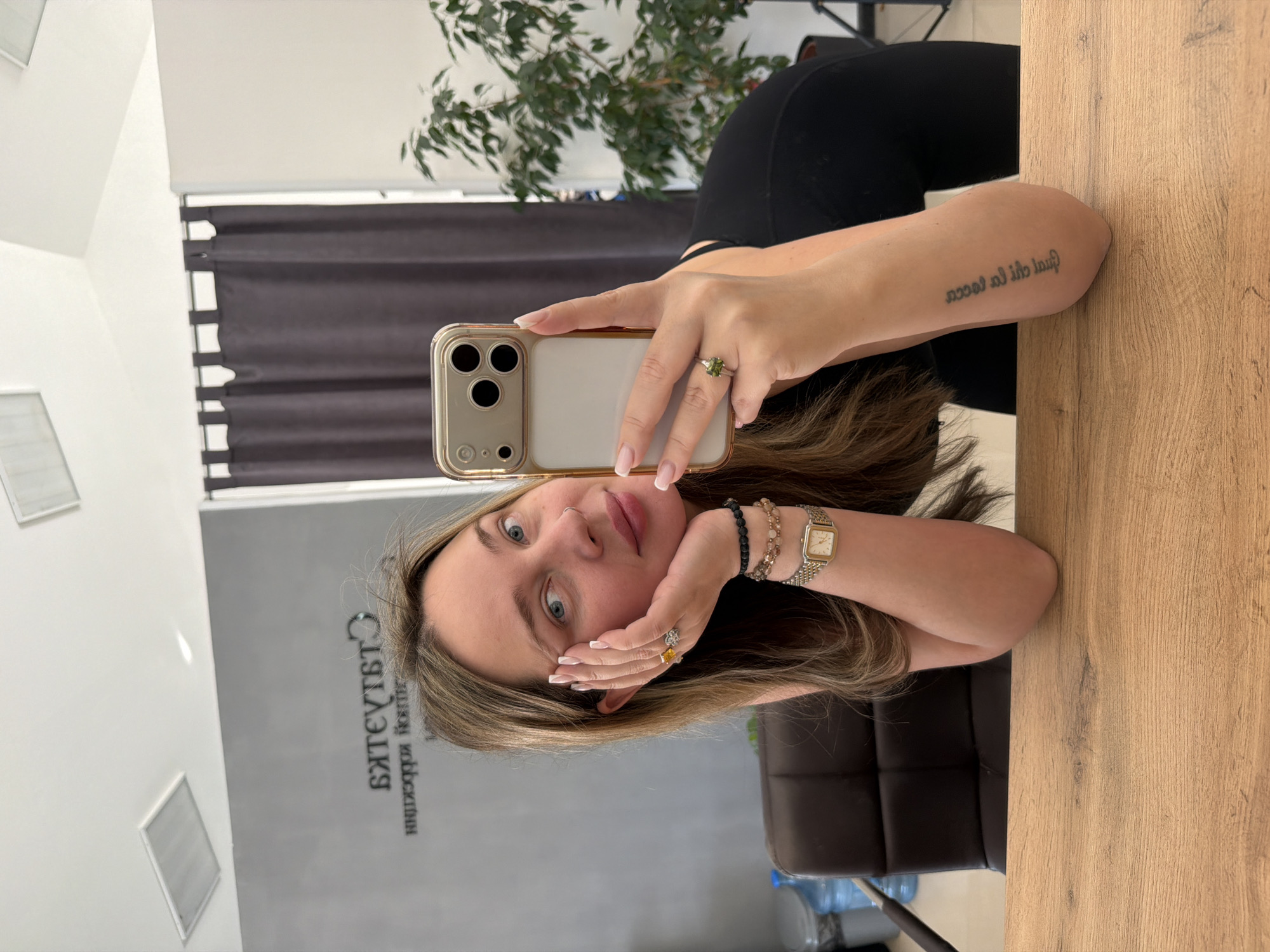 A woman is taking a selfie in front of a mirror. She is holding a gold iPhone in her right hand. She has fair skin, blue eyes, and blonde hair. She is wearing a black shirt, and several rings, bracelets, and a watch. The mirror reflects the interior of a room with a grey wall with the writing 'BATE/TE/TA' in Russian in Chaikovsky, Russia. There is also a black curtain, a plant, and a wooden table in the room.