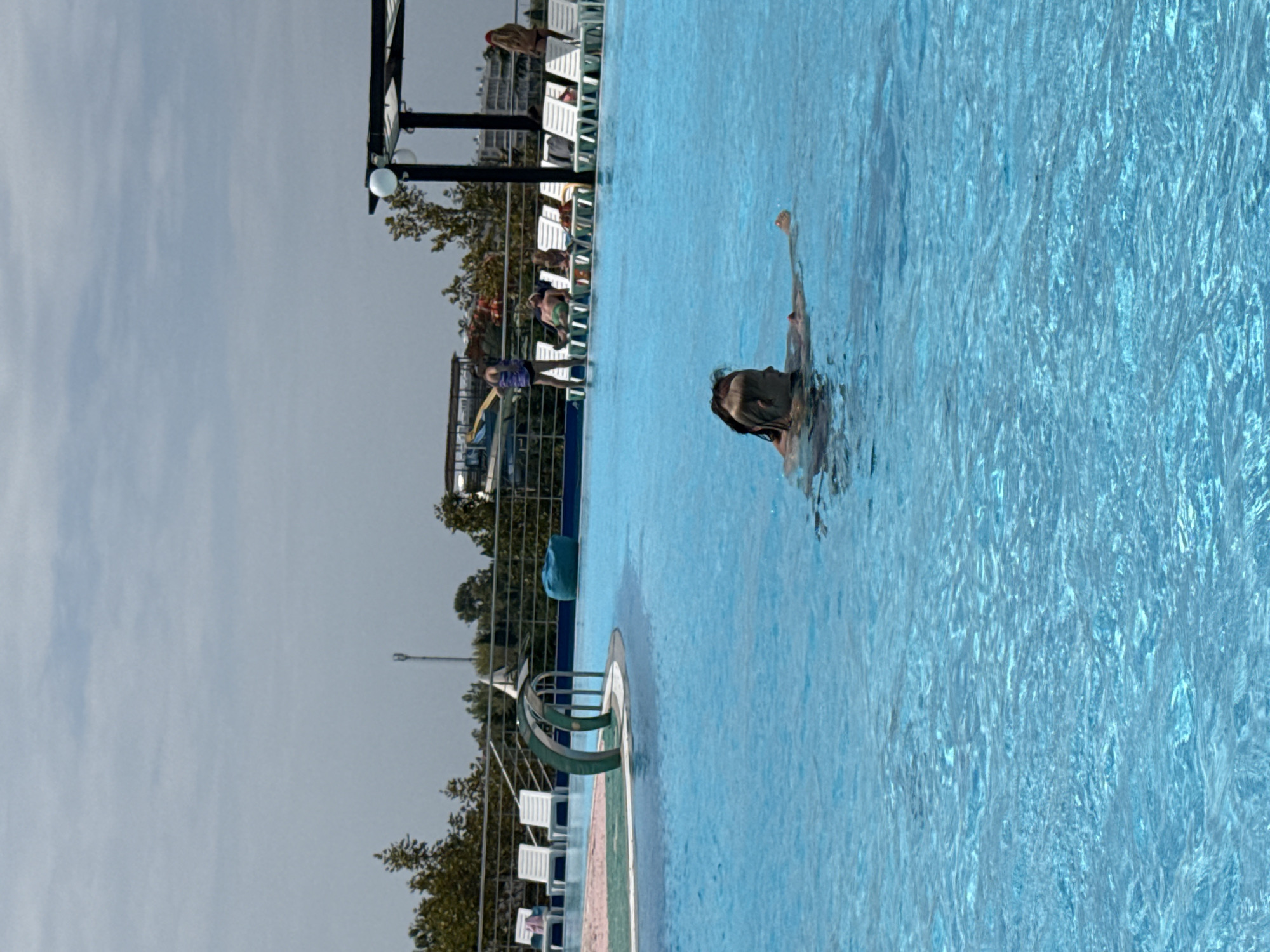The image shows a person swimming in a bright blue swimming pool. The person is partially submerged in the water, with their head and shoulders visible. They appear to be wearing goggles. In the background, there are lounge chairs, trees, and a structure that could be part of a water park. The sky is overcast. There is no information to indicate the location is in Sevastopol, Ukraine.