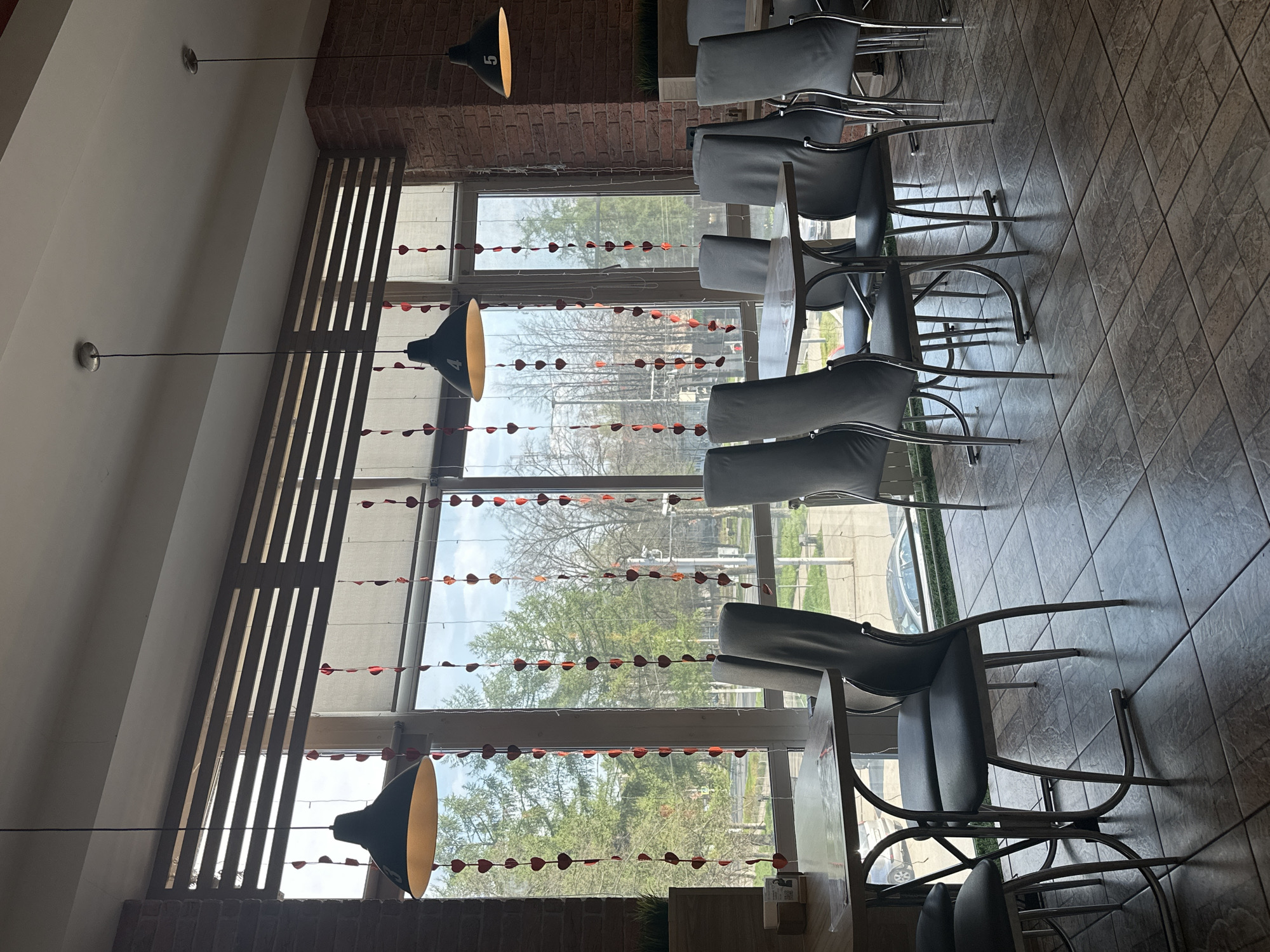 The image shows the interior of a building, possibly a restaurant or cafe, located in Chaikovsky, Russia. The scene includes tables and chairs arranged near large windows decorated with heart-shaped garlands. Outside the windows, trees and buildings can be seen, suggesting an urban or suburban environment. The interior is lit by hanging lamps, and the overall atmosphere is one of a public space prepared for customers. There are no people visible in the frame. The focus is on the furniture and the view through the windows.
