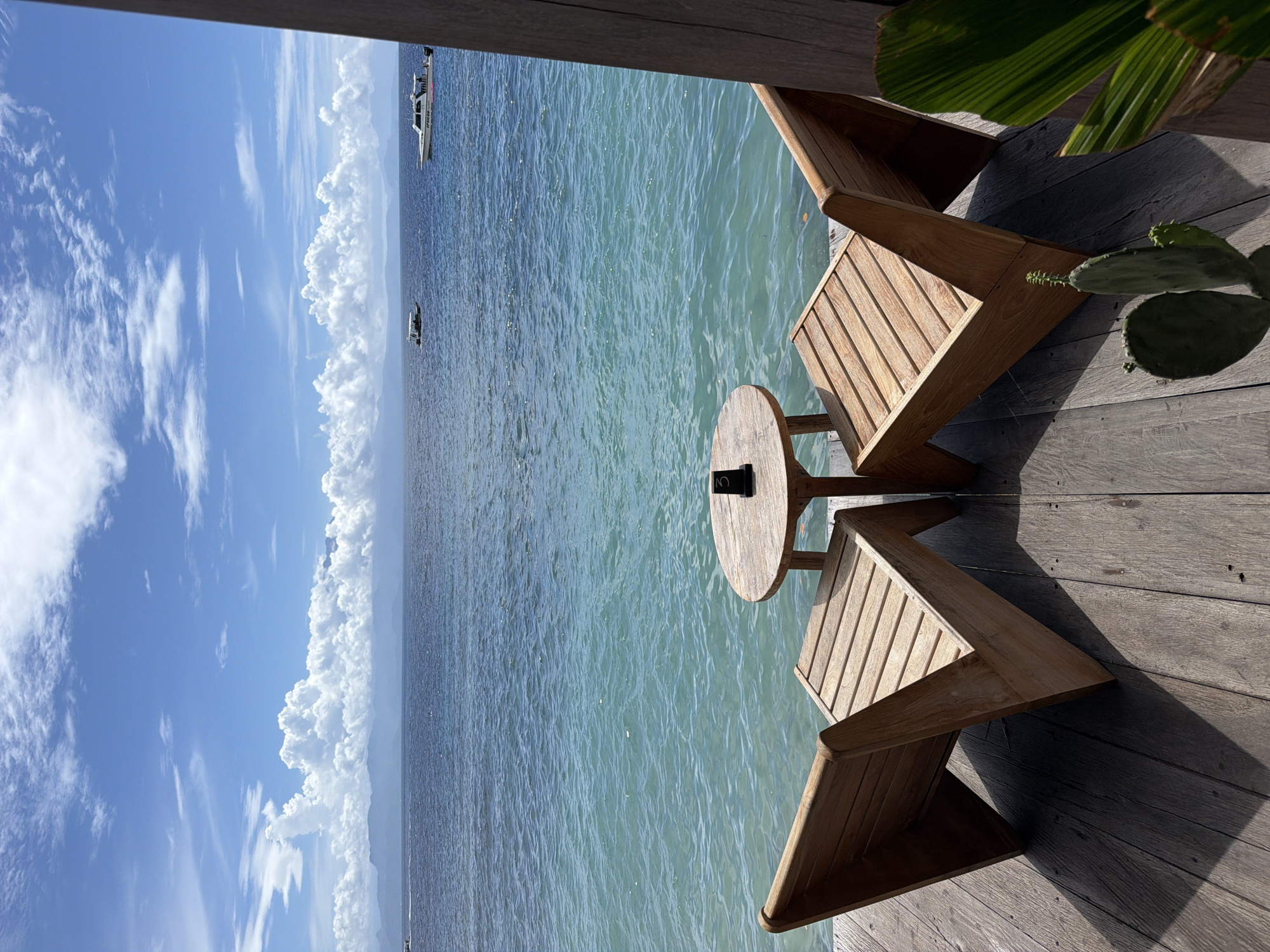 The image shows a serene scene at a coastal spot likely in Nusa Penida, Indonesia. The perspective is slightly elevated, capturing a wooden deck with two wooden chairs and a small round table. The table has a sign that reads "3." In the distance, the ocean stretches out, its surface a mix of blue and turquoise hues, reflecting the sky. Two boats are visible on the water. Beyond the water is a bank of puffy white clouds with a clear blue sky above. A part of a cactus is visible in the top-right of the image.