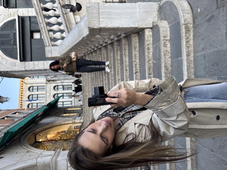 The image depicts an outdoor urban scene in Venice, Italy, specifically on or near a major stone staircase associated with a bridge, characteristic of Venetian architecture.
In the foreground, positioned to the left and slightly angled, a young adult female with long brown hair, wearing a beige trench coat over a plaid shirt and light blue jeans, holds a black digital camera with both hands, looking down at its screen or viewfinder as if composing a photograph. A silver-toned ring is visible on her right ring finger.
In the mid-ground, an adult female with dark curly hair, dressed in a dark olive-green jacket, dark trousers, and white sneakers, descends the stone staircase. She holds a mobile phone in her right hand, looking at its screen, and carries a light brown shoulder bag on her left side.
The environment features a wide, ancient stone staircase with a balustrade on the right. To the left, a stone archway reveals a shop entrance with internal lighting and a green awning above. Beyond the mid-ground woman, other indistinct pedestrians are visible on the steps. In the background, classic Venetian buildings with multiple arched windows and dark shutters are present under a clear sky. A dark pigeon is captured in mid-flight on the right side of the image, near the stone balustrade. The ground is paved with stone slabs.