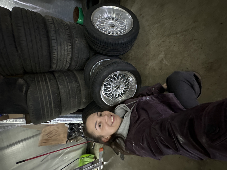 An indoor scene in Zhabkino, Russia, depicts a female subject posing in a garage or storage area surrounded by vehicle tires. The woman, with dark hair pulled back, is positioned in the foreground, crouching or kneeling, facing the camera with a smile. She wears a grey hooded sweatshirt under a dark purple jacket and dark trousers. Behind her, two tall stacks of dark-colored used tires reach towards the ceiling. To the right of the woman, four new or recently cleaned tire-and-wheel assemblies are arranged on the concrete floor, featuring black tires mounted on multi-spoke silver alloy wheels. The background walls are covered with a reflective material. To the far left, a red-handled tool leans against the wall, next to a partially visible metallic step ladder and a bright green bucket on the floor.