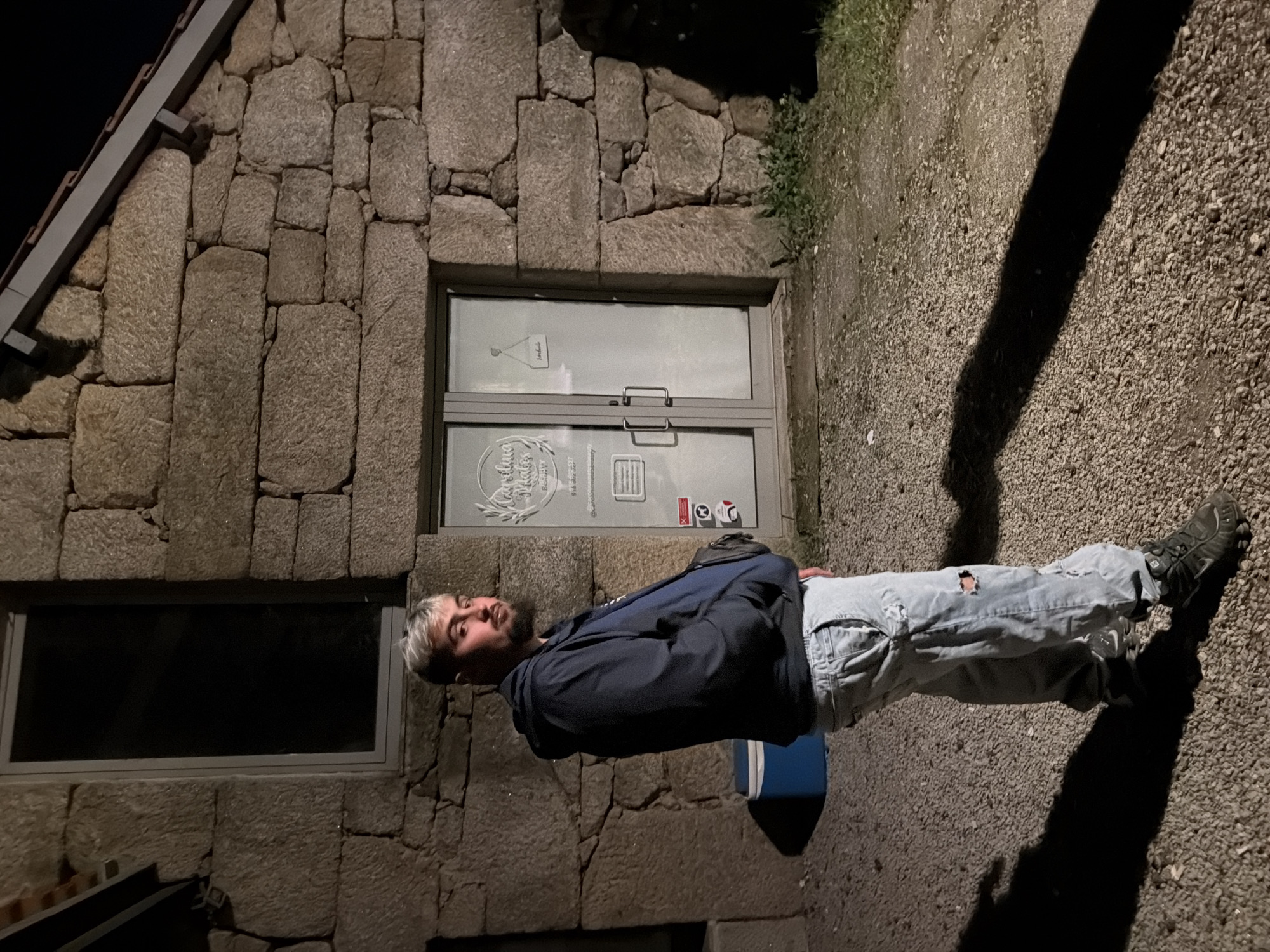 A man stands in front of a stone building with a window and a glass door. He wears a dark jacket, light-colored ripped jeans, and dark shoes. The ground is gravel. The building is likely located in Vila Real, Portugal.
