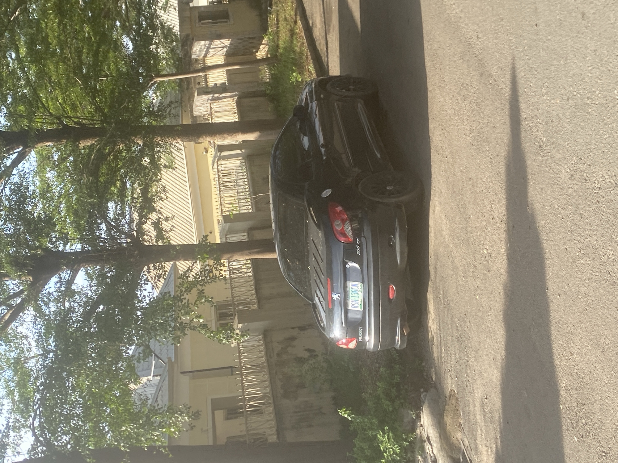 A black Peugeot 206 CC is parked on the side of a street in an urban environment. The car is positioned near a curb with a building in the background, partially obscured by trees. The building has a light-colored exterior with decorative metal railings. The street is paved, and there are some shadows visible on the surface. A license plate reading "RSH-136CN" is visible on the back of the car. The location is Unknown, Unknown.