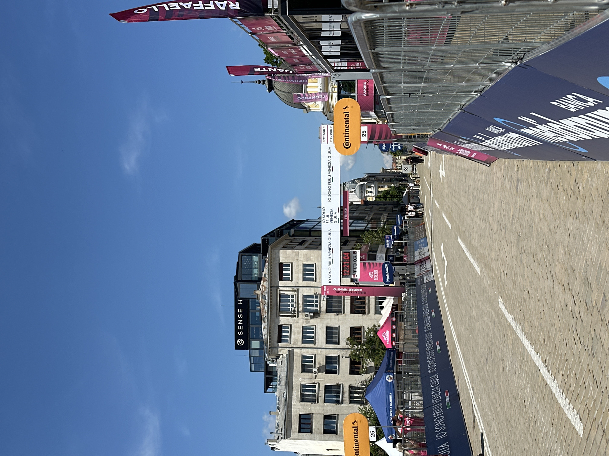 This image depicts a street scene in Sofia, Bulgaria, likely during a cycling event. The road is paved with cobblestones and marked with white lines, suggesting it is a designated course. Barriers are set up along the sides, with signage promoting various sponsors like Continental, Tudor, Giro d'Italia and Banca. There are also several banners with the logos of Giro d'Italia. On the left, buildings are visible, including one that has "SENSE H" on it. The sky is clear and blue. There are people in the background behind tents and other event-related infrastructure. The overall impression is of a street set up for a race, with the focus on the infrastructure and the lack of active participants.