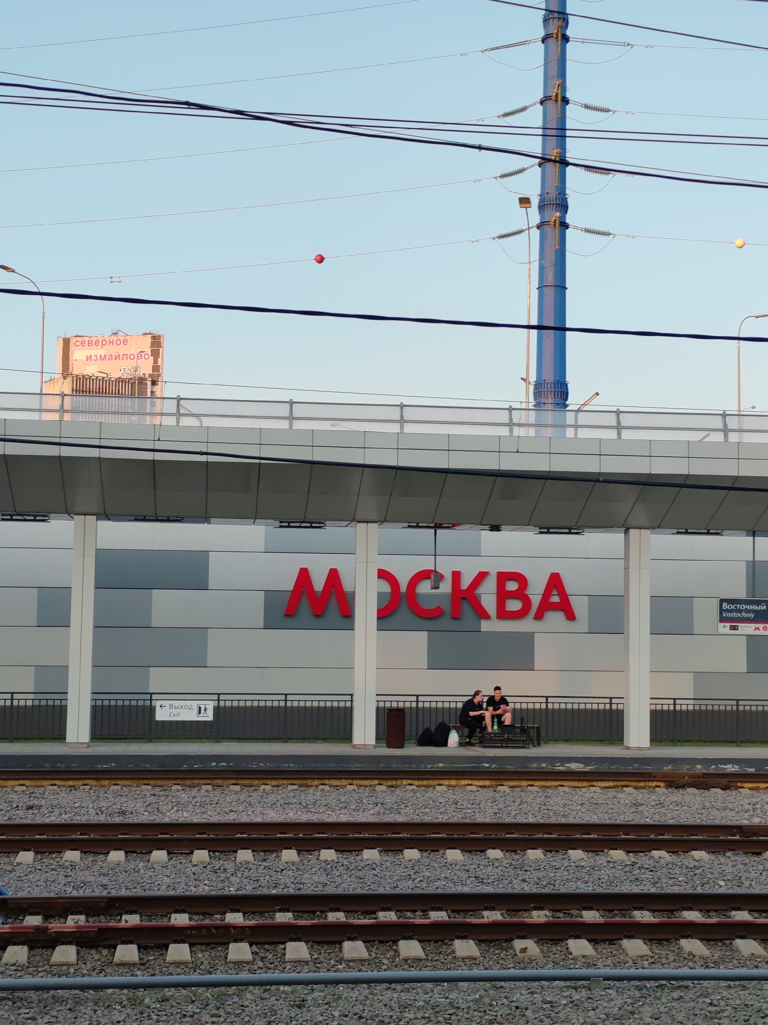 In Moscow, Russia, two young people sit on a bench at a train station platform. They appear to be a couple, one looking at their phone while the other drinks from a bottle. The station platform features a modern design with large, grey, rectangular tiles on the walls. Prominently displayed on the wall is the word "МОСКВА" in large red letters, indicating the station's location. To the right, a smaller sign reads "Восточный Vostochniy" with a Metro logo and station indicator. An "Exit" sign with an arrow pointing left and a pictogram of a person walking is visible on the platform wall.
The scene is set against a backdrop of a clear, light blue sky, suggesting it is likely daytime, possibly late afternoon or early evening given the warm lighting on the building. Overhead, thick power lines stretch across the frame, supported by a tall, blue, lattice-like pylon. In the distance, a large building with a sign reading "северное измайлово" (Severnoye Izmaylovo) is visible, further contextualizing the location. Railway tracks, filled with grey gravel, run in the foreground, separating the viewer from the platform. The overall atmosphere is calm and serene, with the two individuals appearing relaxed while waiting.