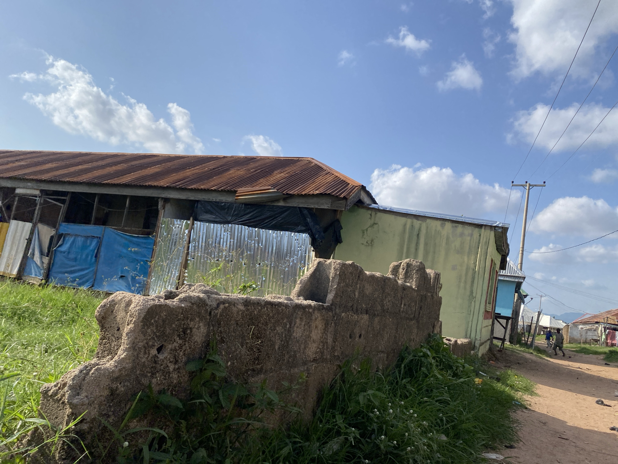 A wide shot captures a rural Nigerian street scene under a partly cloudy sky, suggesting a mid-day setting. The foreground is dominated by a weathered, rough-hewn concrete wall, overgrown with lush green grass and small white flowers. To the left, a long, rustic building with a corrugated tin roof, showing signs of rust, stands partially obscured by the wall. This structure appears to be constructed with a mix of materials, including corrugated metal siding and blue tarpaulin sheeting.
The scene extends down a dusty, unpaved road that curves towards the background. On the right side of the road, more modest dwellings with tin roofs are visible, some with washing hanging outside. A utility pole with several wires stretches across the frame, indicating the presence of electricity. In the distance, two figures, likely men, walk along the road, one in a blue shirt and the other in a camouflage-patterned outfit. The overall impression is one of a lived-in, informal settlement, typical of many rural areas in Nigeria. The weather appears clear with scattered white clouds against a bright blue sky. There is no visible text in the image.