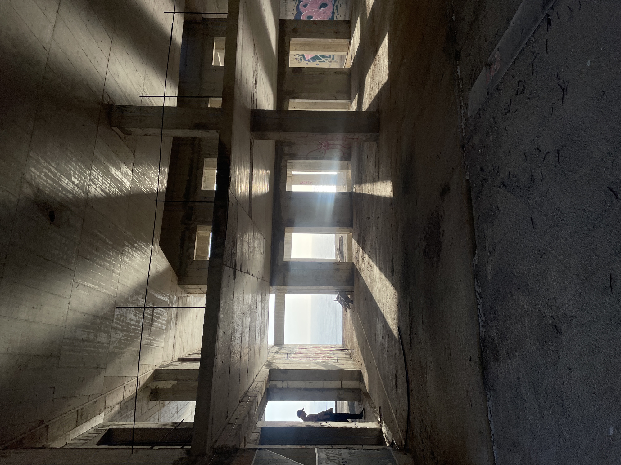 The image depicts the interior of an unfinished or abandoned concrete structure, possibly located in Donji Katun, Montenegro, viewed from a tilted, low-angle perspective, looking upwards. The structure features repetitive, rectangular openings that admit sunlight. One person lies prone in the lowest opening. The concrete walls exhibit varied light and shadow patterns. Graffiti is visible on some of the higher wall sections. Metal reinforcement rods extend from the concrete in places.