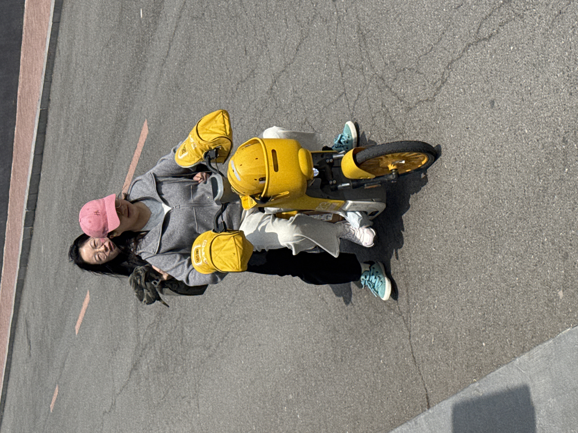 The image, best viewed rotated 90 degrees counter-clockwise, depicts two individuals, likely women, resting outdoors next to a prominent bright yellow electric scooter on an asphalt surface under clear daylight.
One woman, wearing a grey zip-up hoodie, light-colored pants, and a pink baseball cap, is seated or standing upright beside the scooter. She has dark hair and is wearing glasses. The second woman, with dark hair and a dark top, is leaning against the first woman's back and shoulder, appearing to be in a relaxed state with her eyes closed or looking down, suggesting a moment of rest or comfort.
The central object is a distinctive yellow electric scooter or bicycle, featuring a yellow front basket or compartment. Two yellow insulated bags, often used for food delivery, are attached to the scooter; one is clearly visible on the handlebars. The scooter has black tires with yellow rims. Both women are wearing sneakers: one has light blue/teal shoes, while the other wears white or light pink sneakers.
The setting is an outdoor paved area, likely a street or pathway, characterized by dark asphalt with minor cracks and faint road markings, including a thin orange-pink line and a white line. A raised curb or sidewalk is visible on the left side of the frame. The presence of strong shadows indicates a sunny day. The scene captures a casual break, possibly during a delivery route or leisure ride, in Zhongtun, China.
Notable details include:
- The pink cap worn by one woman features a logo resembling a stylized 'H'.
- On one of the yellow insulated bags, the Chinese characters "美团" (Meituan) are clearly visible, identifying it as equipment from a major Chinese food delivery platform.
- Faint text or numbers, possibly "E9" or "EQ", can be seen on the side frame of the yellow scooter.