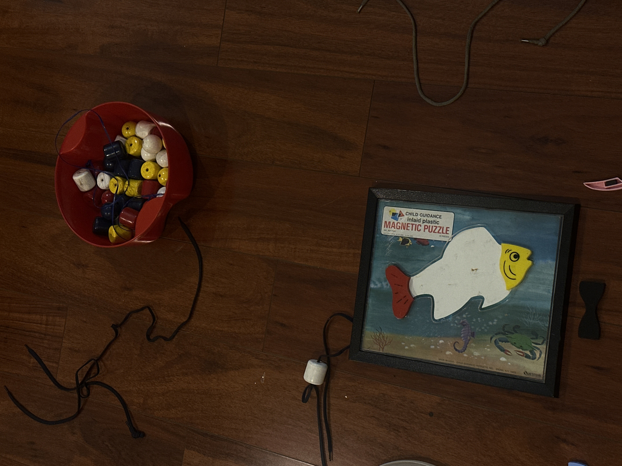 The image shows a collection of toys on a wooden floor. On the left, there's a red plastic bowl filled with colorful wooden beads. Black string is coming out from the bowl. To the right, a "Child Guidance Magnetic Puzzle" depicting a fish in a blue background. Beside the puzzle a black bow tie shape. A gray string appears in the upper right part of the image. The location is not discernible from the provided image. There's no evidence indicating the location is in Huixquilucan de Degollado, Mexico.