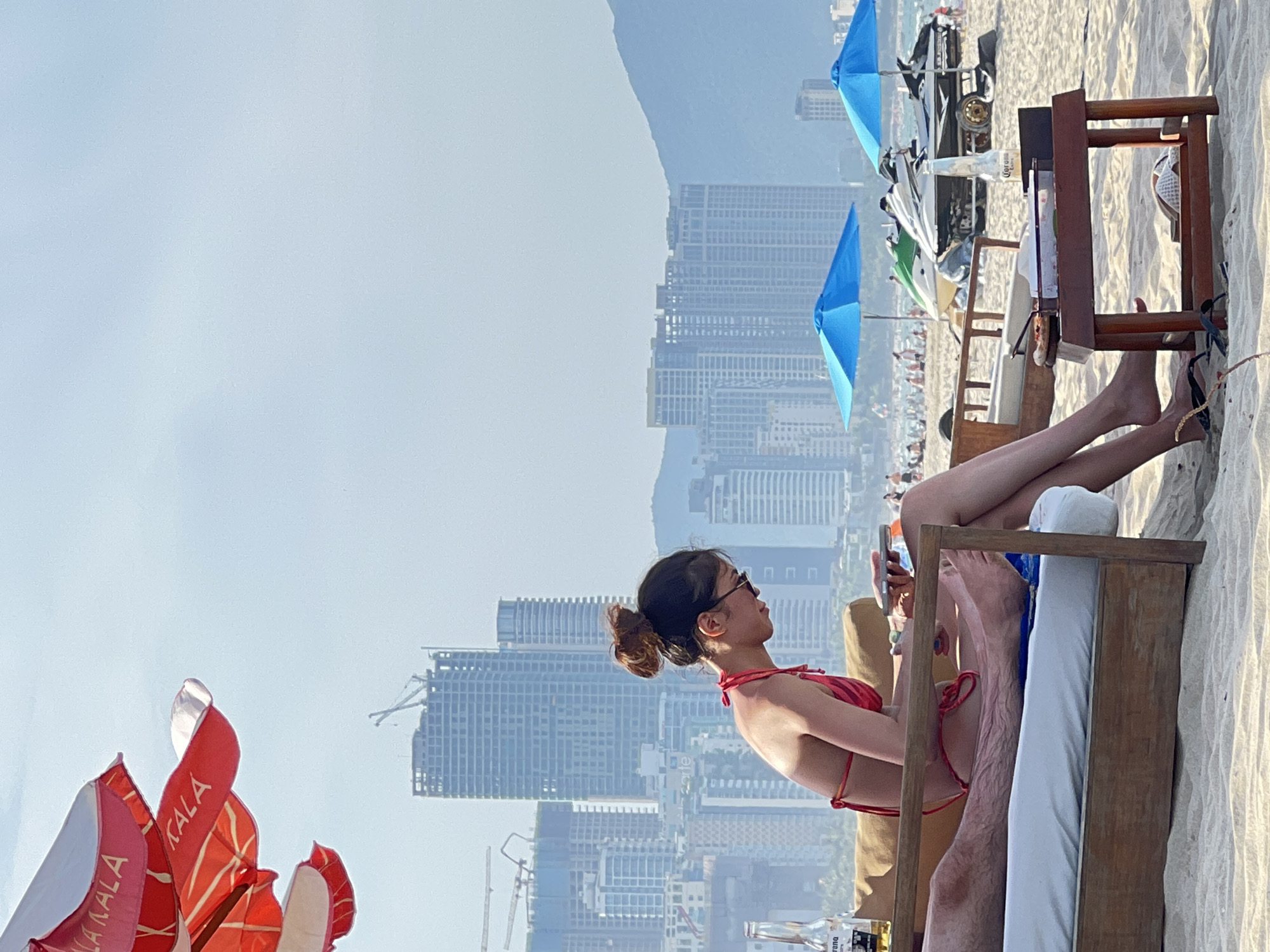 A woman in a red bikini is sitting on a lounge chair on a sandy beach, looking at her phone. She has her hair pulled up in a bun and is wearing sunglasses. There are several buildings under construction in the background, along with some tall apartment buildings and a mountain range. Two blue umbrellas and two jet skis are also visible on the beach. A bottle of Corona beer and a small table with food are next to the woman. This scene appears to be on My Khe Beach in Da Nang, Vietnam, known for its sandy shores, developed coastline with high-rise buildings, and proximity to construction projects.