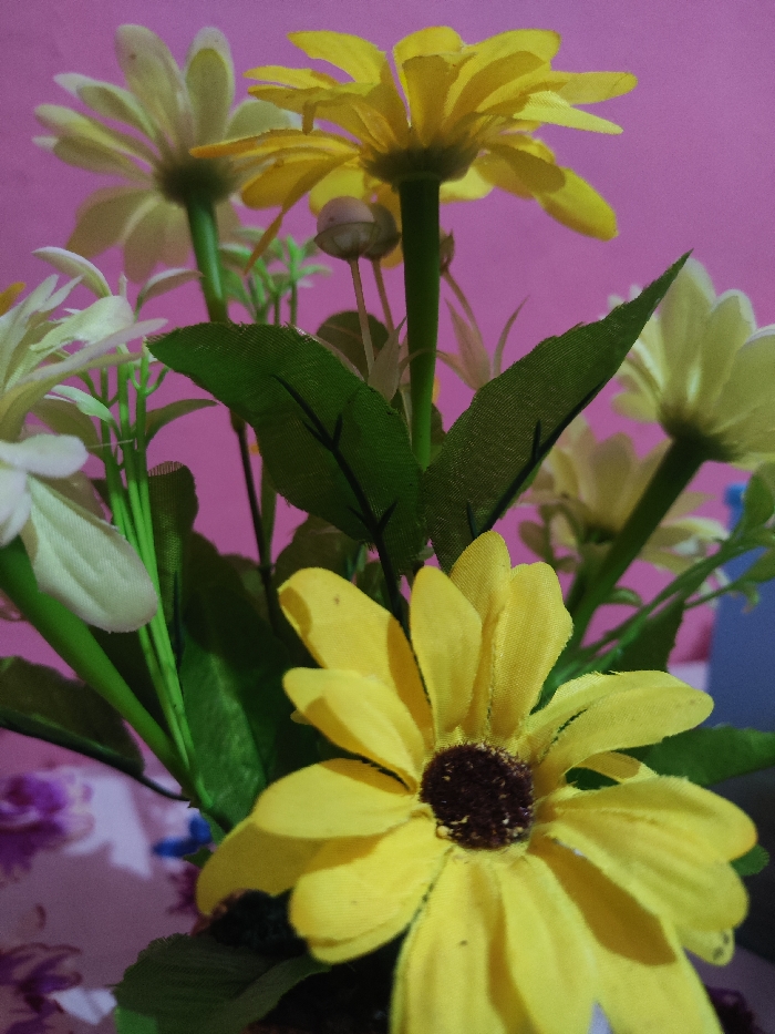 The image displays a close-up view of a bouquet of artificial flowers, situated in a domestic or indoor setting in Astanajapura, Indonesia. The arrangement primarily features daisy-like blossoms in vibrant yellow and pale cream or off-white, constructed from fabric petals. The yellow flowers have distinct dark brown centers. Green artificial stems and leaves are visible, intertwining among the flower heads. The background is a solid magenta or deep pink, likely a wall. In the lower left foreground, a patterned surface, possibly fabric or a tablecloth, displays a muted floral or abstract design in shades of purple and white. The lighting highlights the texture of the fabric petals, and a shallow depth of field keeps the nearest yellow flower in sharp focus while elements in the background are softly blurred. No individuals are present, and no actions or interactions are depicted.