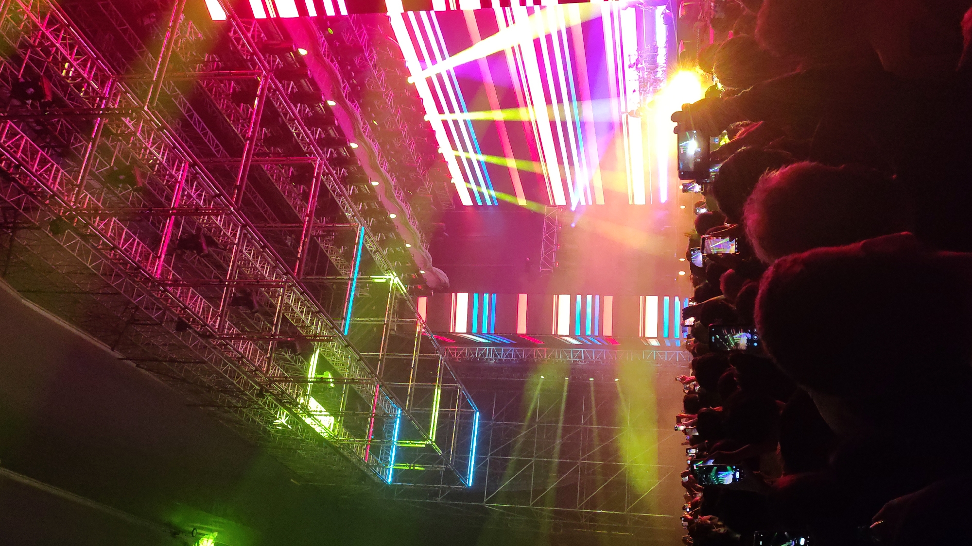 The image depicts a concert or performance scene, likely indoors, taken from the perspective of someone in the audience. The foreground is dominated by the silhouetted heads and hands of audience members, many holding up smartphones to record the event. Beyond the audience, a complex metal structure serves as a framework for lighting and visual displays. The structure is illuminated in vibrant colors, including pink, purple, green, blue, and yellow. Various light beams and panels add to the visual spectacle. Based on the lighting and set-up, the scene appears to be an electronic music event or a similar type of performance. There is no definitive information in the image to identify its location as San Miguel, Peru.