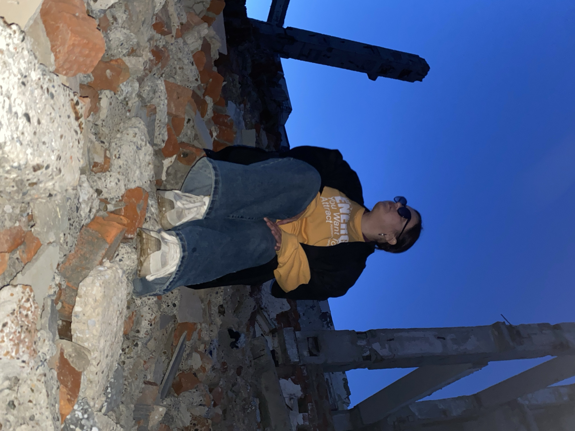 A woman sits on a pile of rubble and debris in what appears to be the ruins of a building or structure. The surrounding environment consists of broken bricks, concrete chunks, and skeletal remains of a building framework, suggesting a site of destruction or demolition. The sky is a deep blue, likely indicating twilight or early evening. The woman is facing upwards, with her gaze directed towards the sky or the upper part of the ruins. She is wearing sunglasses, a yellow top with text, jeans, a dark jacket, and light-colored sneakers. The location is Марковский, Russia.