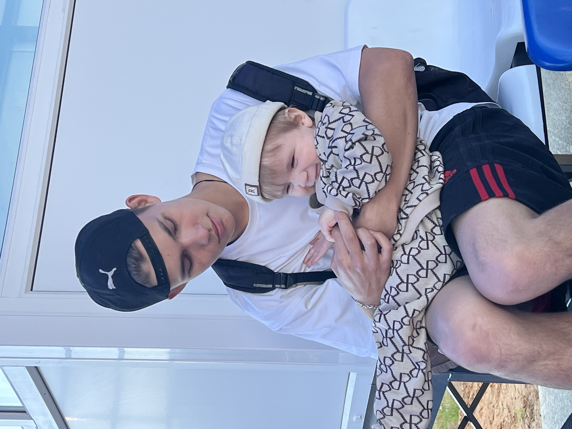 The image captures an outdoor scene in the city of Noviy, Russia, featuring a young adult male holding an infant. The male, appearing to be in his late teens or early twenties, is seated, looking downwards at the infant. He has light skin and wears a black baseball cap backward, displaying a white Puma logo on the rear. His attire consists of a plain white short-sleeved t-shirt, black athletic shorts with three distinct red stripes vertically oriented on the outer left thigh, and a black backpack worn with straps over both shoulders. A silver-colored chain bracelet is present on his left wrist. His knees exhibit visible marks or abrasions.
The infant, held by the male, appears to be between six and eighteen months old, possessing light skin and light-colored hair. The infant wears a light beige beanie with a small red square patch featuring a white "R" on the front, and a one-piece outfit characterized by a repeating pattern of dark geometric shapes on a light beige background. The infant displays a wide smile or laugh, with its gaze directed towards the viewer's left. The male's hands are positioned near the infant's hands.
The background consists of a light-colored, uniform building or modular structure. A white seating surface is visible behind the male's lower body, and a blue object is partially present in the bottom right corner. The ground in the bottom left portion of the image is exposed dirt with minimal green vegetation. The scene is illuminated by bright, natural daylight.