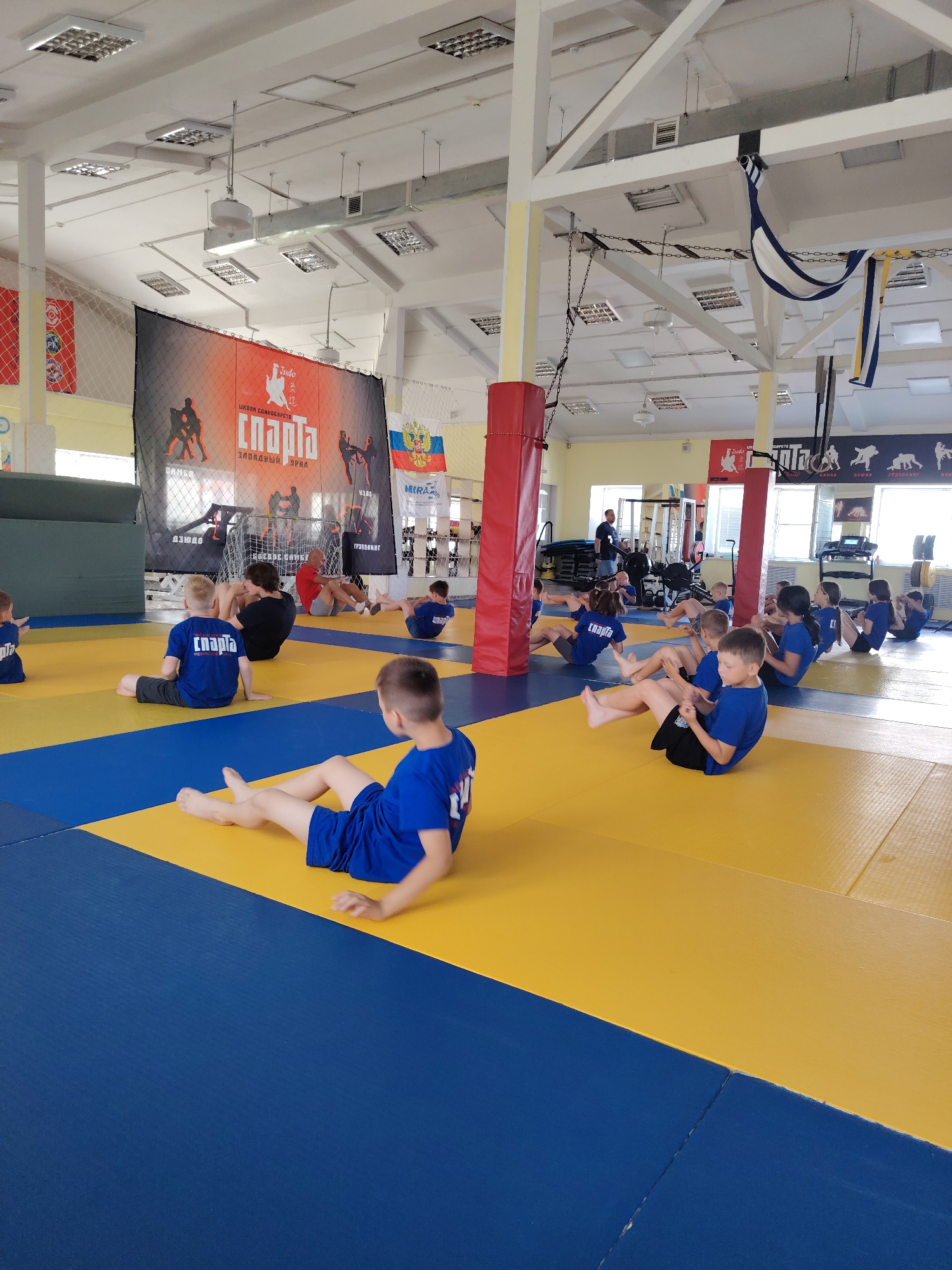 In Chaikovsky, Russia, a group of children, likely participating in a judo or martial arts class, are engaged in physical exercises on a blue and yellow mat in a gymnasium. They are dressed in blue uniforms with "СПАРТА" (Sparta) printed on them. The children are sitting with their legs extended and appear to be doing crunches or a similar core-strengthening exercise. Some are in mid-motion, with their torsos raised or lowered. The gymnasium has high ceilings with fluorescent lighting, exposed beams, and ventilation systems. A large banner with images of judokas and the word "СПАРТА" is hanging on the back wall, along with several flags, including the Russian flag. Gym equipment, such as weight machines and a treadmill, are visible in the background. The overall environment suggests an active sports training facility.