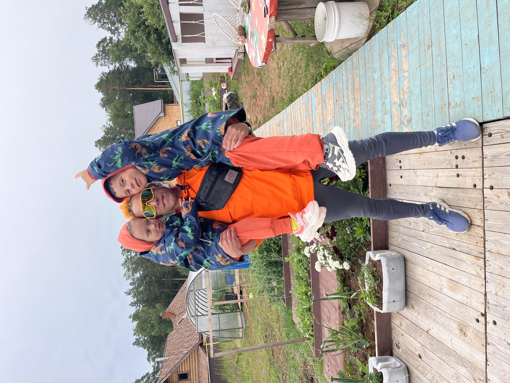 A father is holding two young children in his arms, with one child on his shoulders and the other in front of him. They are all wearing matching blue hooded jackets with a dinosaur pattern. The father is wearing orange pants and a black fanny pack. He has bright yellow sunglasses on. The children are also wearing orange hooded outfits under their jackets. They are standing on a wooden deck, which appears to be in a backyard setting. In the background, there is a house, a greenhouse, and trees. The sky is overcast, suggesting it might be a cloudy day. The scene appears to be a candid moment of family fun and affection. There is no visible text in the image.