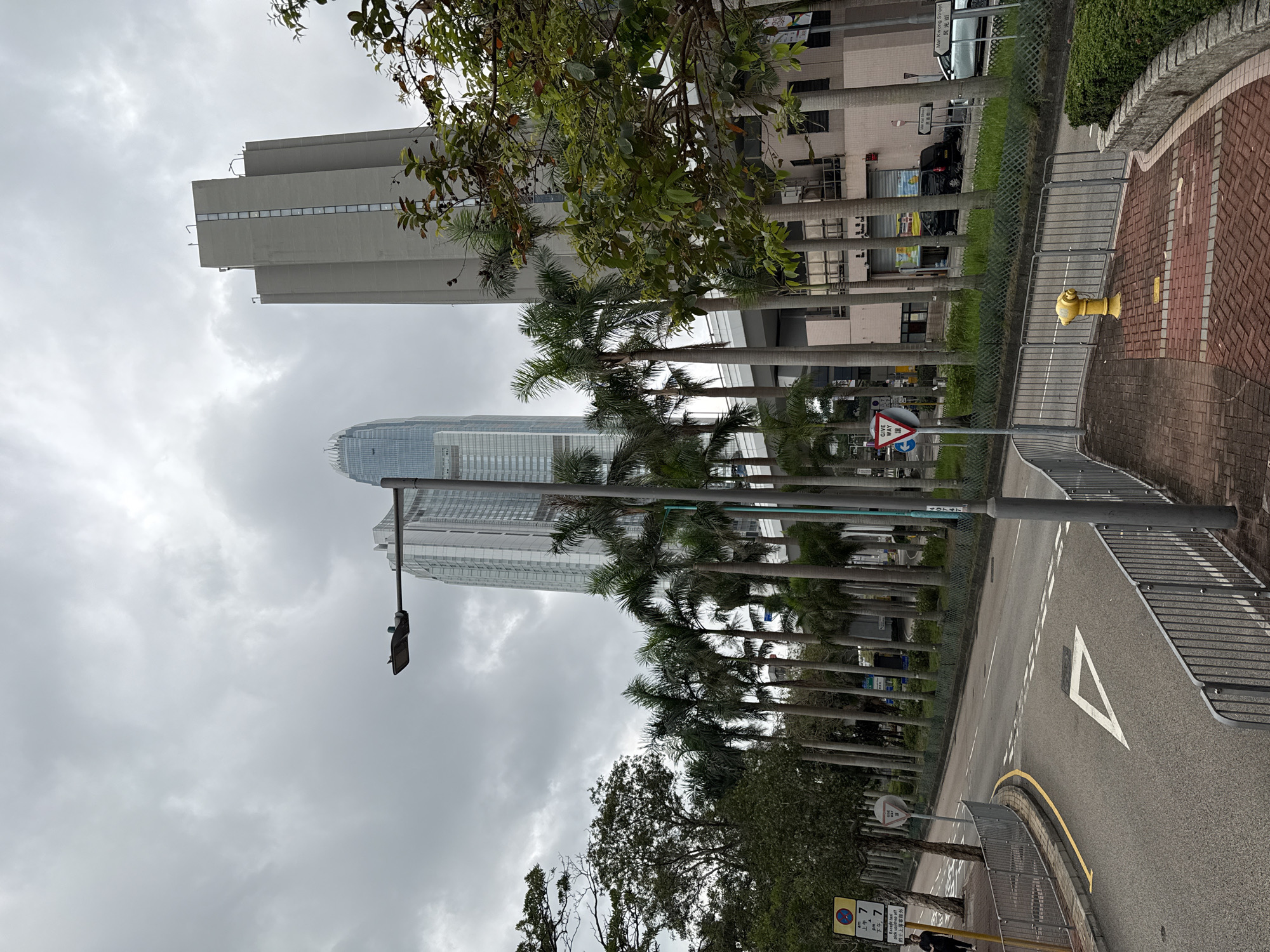 The image depicts a street scene in Hong Kong, China, under a cloudy sky. Tall palm trees line the side of the road, obscuring the view of the skyscrapers in the background, and a row of uniform buildings. On the right side of the image, there is a brick walkway next to a yellow fire hydrant. A white triangular road marking is visible on the asphalt of the road, and a "Give Way" sign can be seen on the left. A metal fence runs along the edge of the road and sidewalk, providing pedestrian safety. A traffic light hangs on the side of the road.