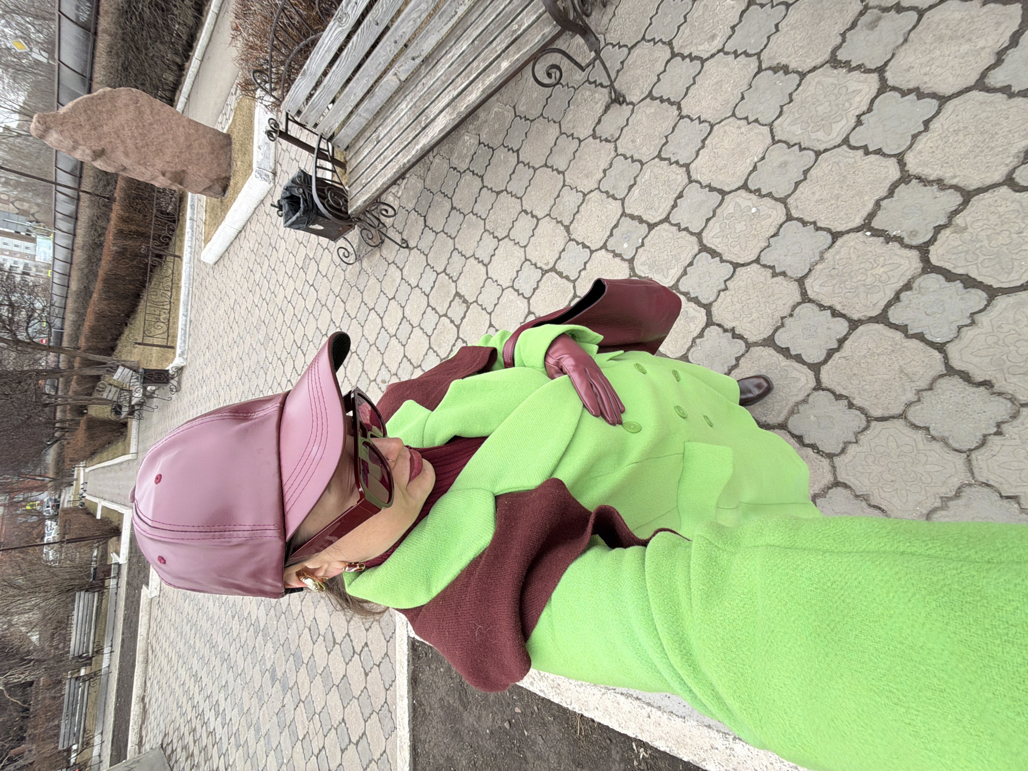 The image is taken in the city of Krasnoyarsk, Russia. It shows a person standing on a sidewalk with a distinctive pattern of light gray paving stones. The person is wearing a bright green coat with brown trim, a matching brown hat, and brown gloves. They also have sunglasses on. The person is standing near a bench with a metal frame and wooden slats. There is a rock sculpture on the sidewalk near the bench. In the background, there are trees and buildings. The overall environment suggests an urban park or public space.