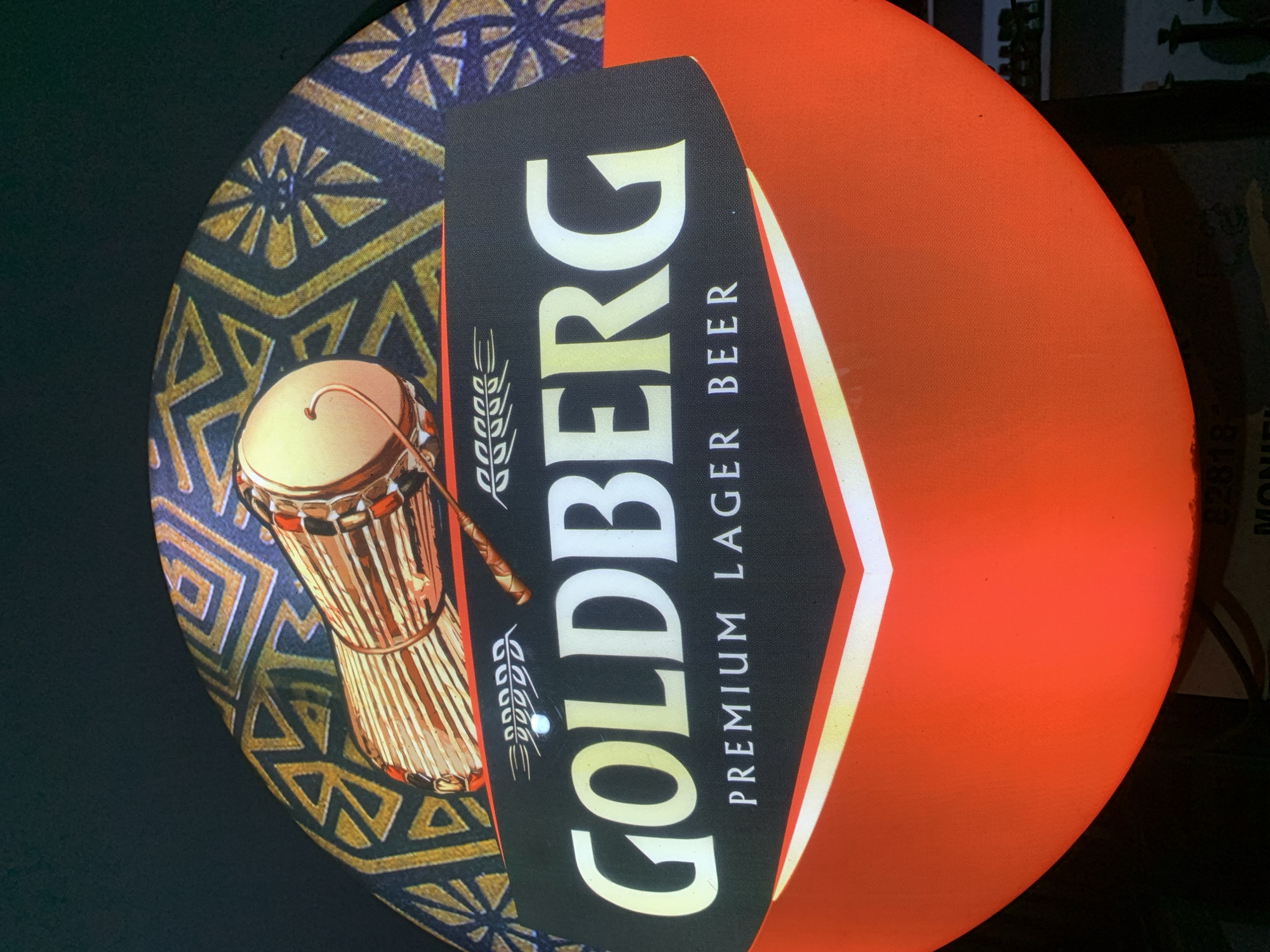 An illuminated, circular sign for "GOLDBERG PREMIUM LAGER BEER" is prominently displayed, likely in a commercial establishment in New Stadium, Nigeria. The sign features a dark blue and gold geometric patterned background in its upper half, upon which a djembe-style drum with a light-colored head and light brown wooden body is depicted horizontally. Below this, a black band contains the brand name "GOLDBERG" in large, light gold and white gradient sans-serif lettering, with "PREMIUM LAGER BEER" written in smaller, white, sans-serif text beneath it. Stylized white barley ears are positioned to the left of "GOLDBERG," and a small white leaf motif is to the right. The bottom half of the sign is a vibrant orange-red, separated from the black band by a sharp white V-shaped line. The sign is brightly lit, contrasting with the dark, out-of-focus background, where faint numbers "82818" and the word "MONIE" or "DANIEL" are barely visible on an obscured object below the sign.