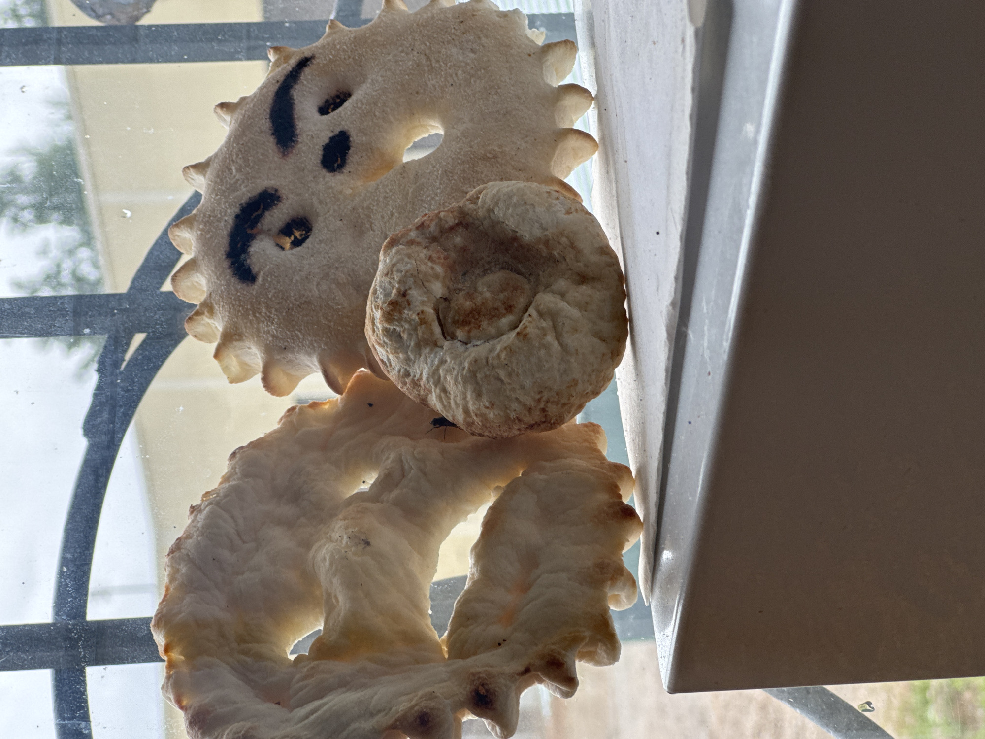 The image displays three pieces of bread-like objects arranged on a reflective surface, likely glass, which is supported by a dark metal structure. The bread pieces have irregular shapes and textured surfaces. One piece, in the upper left, is circular with a serrated edge and has dark markings that resemble a face with two eyes and a mouth. Another piece, positioned above and slightly to the right of the first, is round and dome-shaped with a lighter brown surface. The third piece, at the bottom, is larger and has a more elaborate, twisted shape with a serrated edge similar to the first piece. The lighting suggests it might be daytime, with sunlight casting shadows and highlighting the textures of the bread. The background is out of focus, showing a light-colored wall or structure to the right and a blurry outdoor scene with hints of greenery and possibly a fence or railing to the left. The overall scene is casual and informal, with the bread appearing to be the primary subjects. There is no visible text in the image.