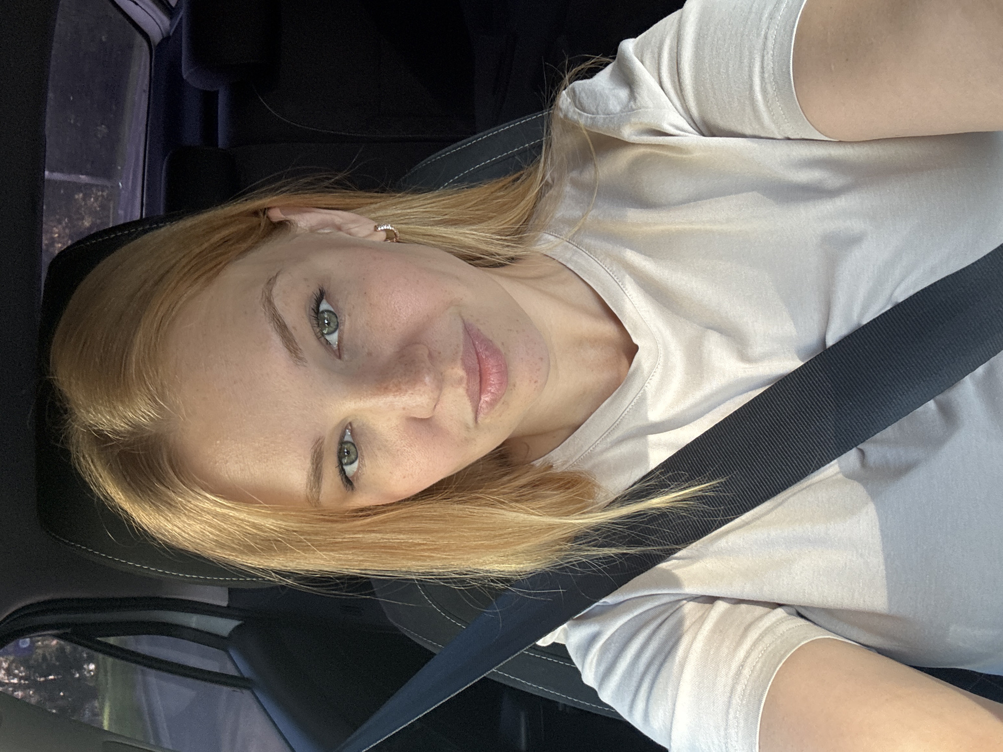 A young woman with blonde hair and freckles is pictured from the chest up, seated in the driver's seat of a car. She is wearing a light-colored t-shirt and a black seatbelt is visible across her chest. Her left arm is partially visible, resting by her side. The background shows the interior of the car, including the headrest and part of the car's interior panel. Outside the window, trees and foliage are visible, suggesting a location in a natural or semi-natural environment. The lighting indicates it is daytime, likely late afternoon or early evening, given the soft light.