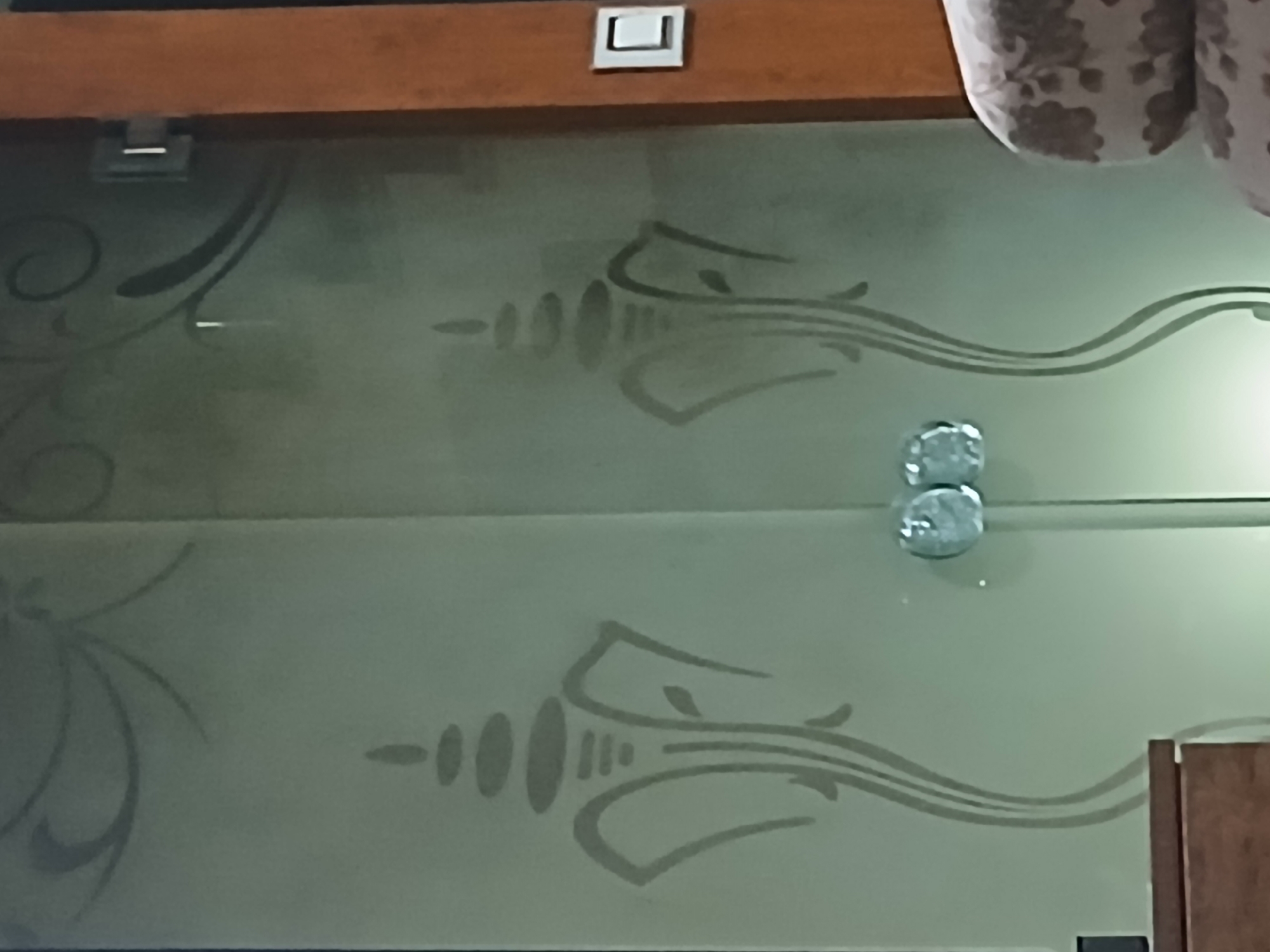 A close-up shot depicts a textured glass surface, possibly a shower door or partition, featuring etched designs of stylized swirls and curvilinear shapes. Two chrome doorknobs are centrally located, positioned one above the other, suggesting a double-door design. A wooden frame or trim is visible at the top of the glass.
The lack of discernible architectural styles or contextual cues prevents the identification of a precise location within Bengaluru, India.