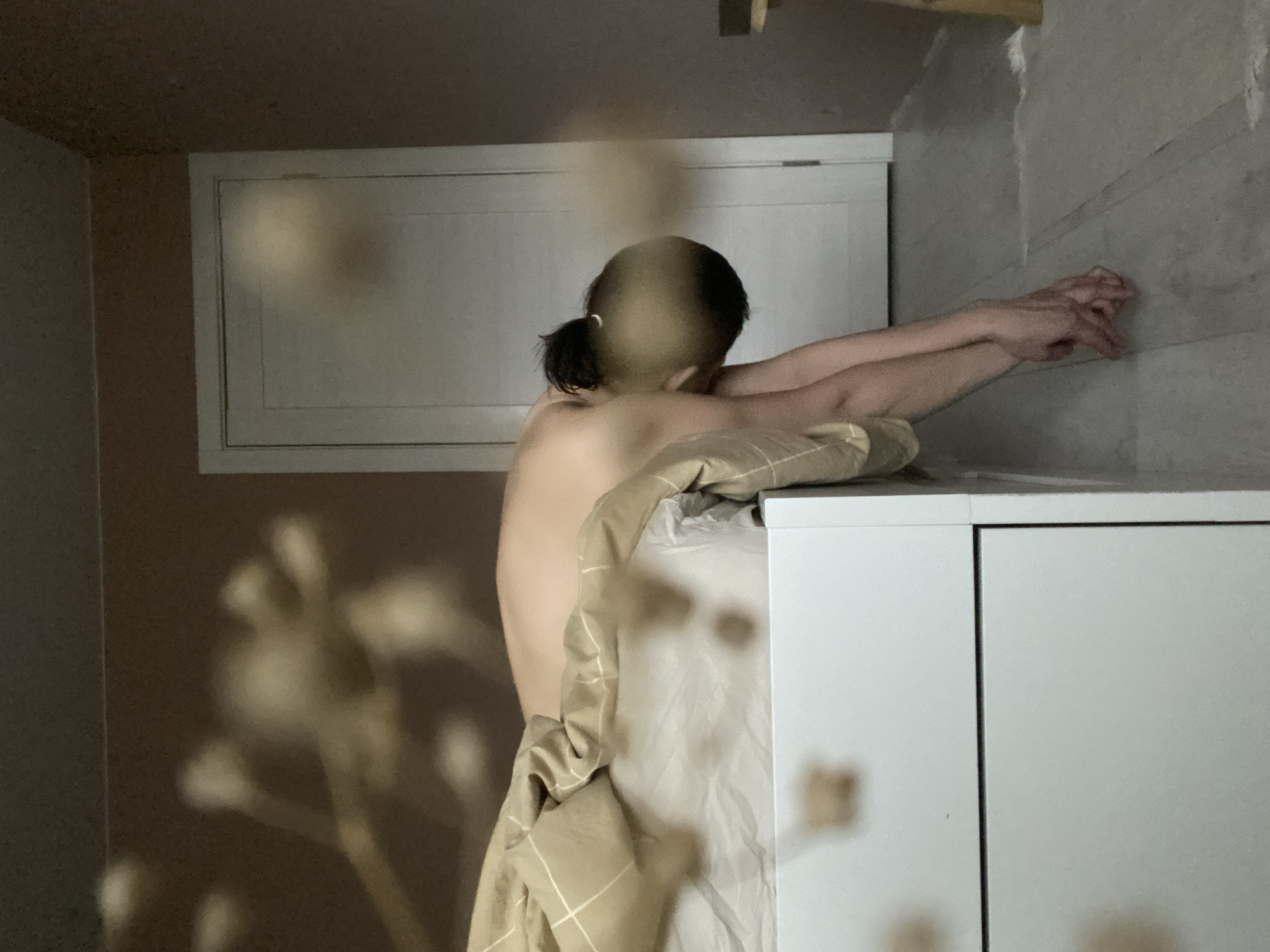 A nude person with short dark hair pulled back in a low ponytail leans against a white cabinet with their arms outstretched, touching a light gray wall. A tan and white plaid blanket drapes over part of the cabinet and the person's body. A white window is visible in the background. Blurred plant stems and heads are in the foreground. The location is Chaikovsky, Russia.