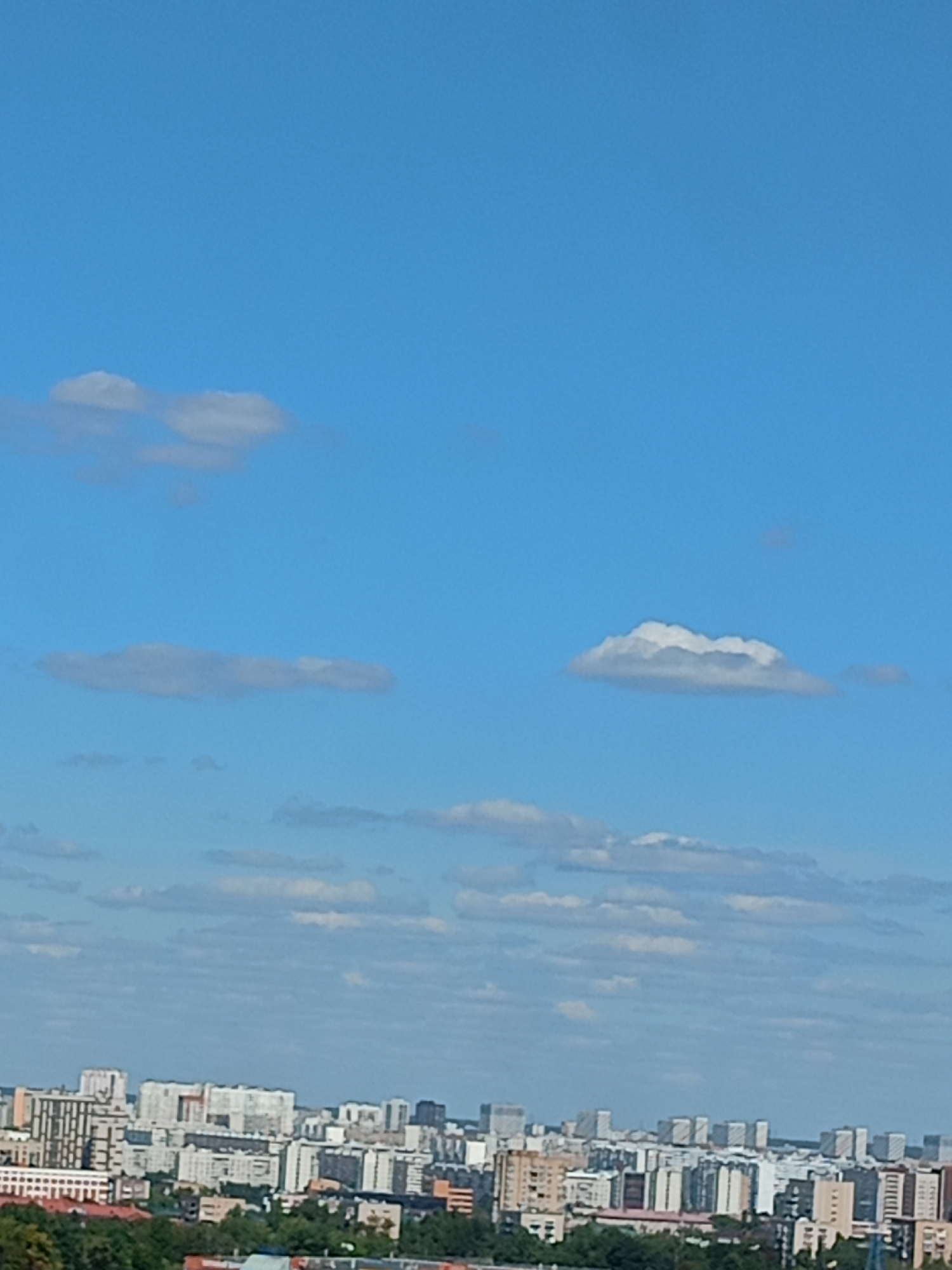 A medium shot captures an urban landscape beneath a bright blue sky dotted with white clouds. The lower portion of the frame is filled with a dense collection of buildings, ranging in height and architectural style, indicative of a city. Trees are visible at the very bottom, suggesting a park or a green space bordering the built-up area. The sky takes up the majority of the image, with several distinct cloud formations scattered across it. The lighting suggests it is daytime, likely mid-morning or afternoon, given the clarity of the sky and the shadows that might be implied by the cloud formations. There are no people or vehicles visible in the image.