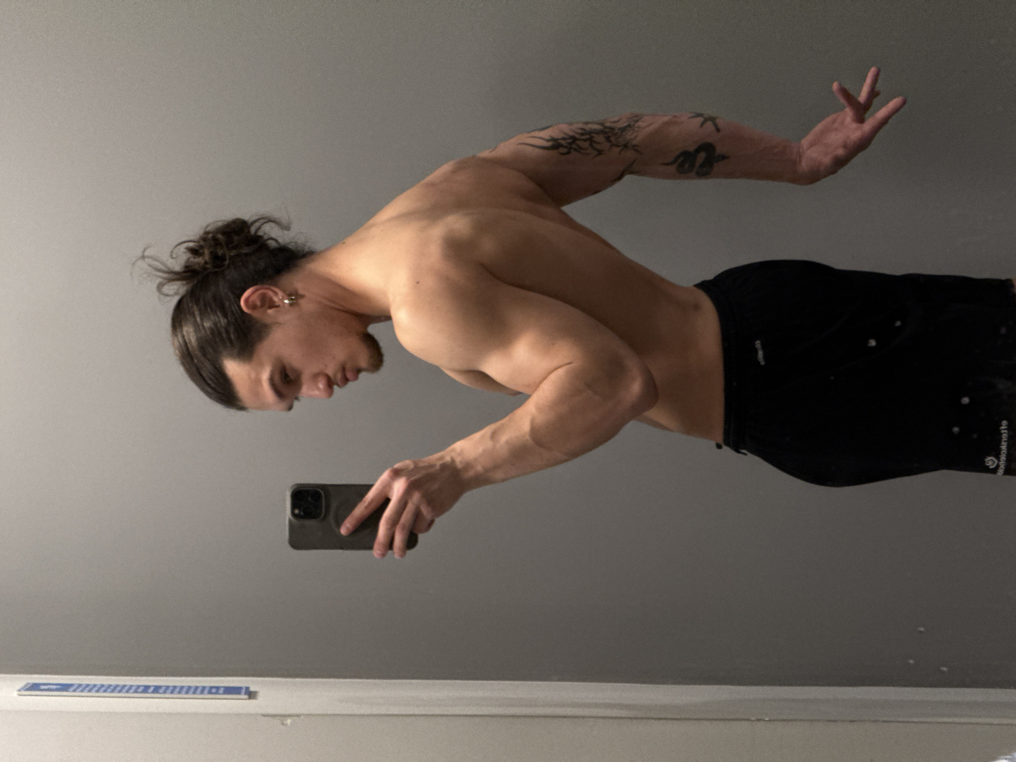 A shirtless man with a bun hairstyle and an earring is standing indoors, holding a cell phone in his left hand, presumably taking a selfie. He is wearing black shorts. He has a tattoo on his upper left arm. The room has gray walls and a white surface below. There is no indication of this image being in Sofia, Bulgaria.