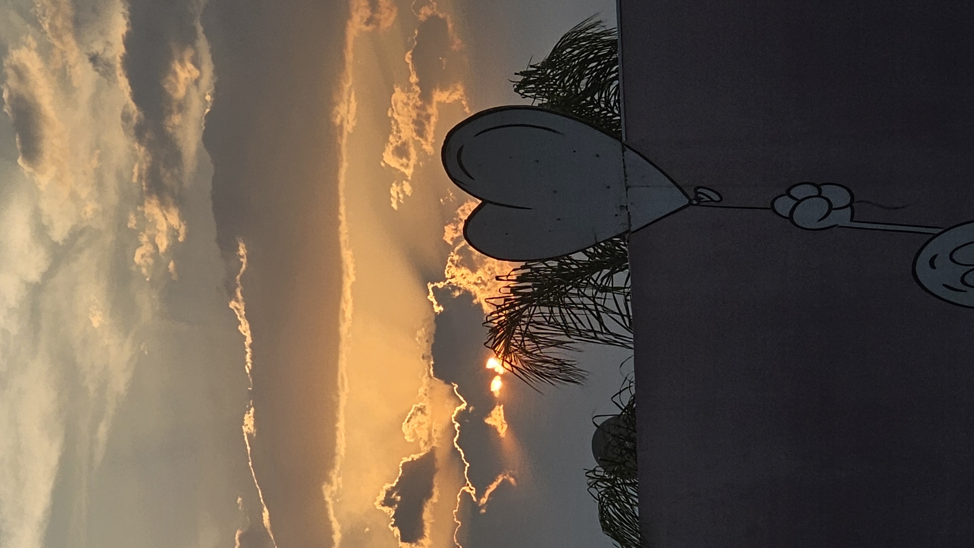 The image depicts a vibrant sunset in Puebla, Mexico. In the foreground is a building with a mural featuring cartoonish, simple line drawings of hearts and other shapes. The colors on the building and the mural are muted due to the angle of the sun. In the background, the sky is filled with clouds and the golden glow of the setting sun. The sunlight filters through the clouds, creating a dramatic effect. Palm trees and other greenery are partially visible between the building and the sky.