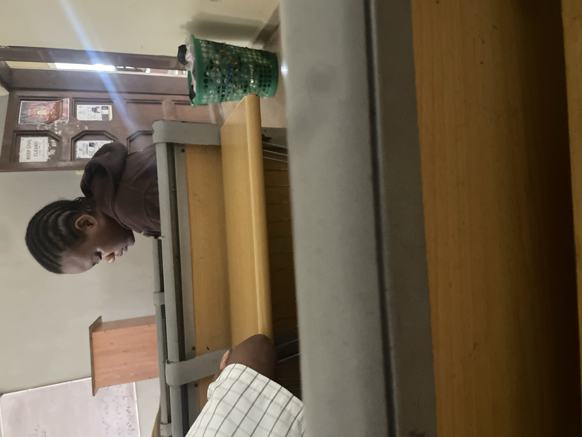 The image shows a student sitting at a desk in a classroom, possibly in Ihiagwa, Nigeria. The student is facing to the side, with their head down, appearing to be studying. They have braided hair and are wearing a brown hoodie. In the background, there is a whiteboard with some writing on it, a wooden box on a stand, a door with various signs and posters, and a green plastic basket filled with items. The classroom appears to be dimly lit.