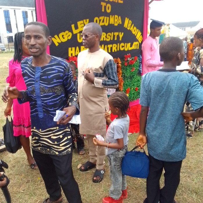 A group of people are outdoors in front of a banner that reads, "Welcome to Kings Ley Ozumba Mbadive University Happy Matriculation". A man in a blue and black patterned shirt stands on the left, holding papers. Beside him is a man in a tan shirt. A young girl in a gray shirt, jeans, and red shoes stands between them, holding a bag. A boy in a blue shirt is on the right with a bag. Other people are visible in the background.
